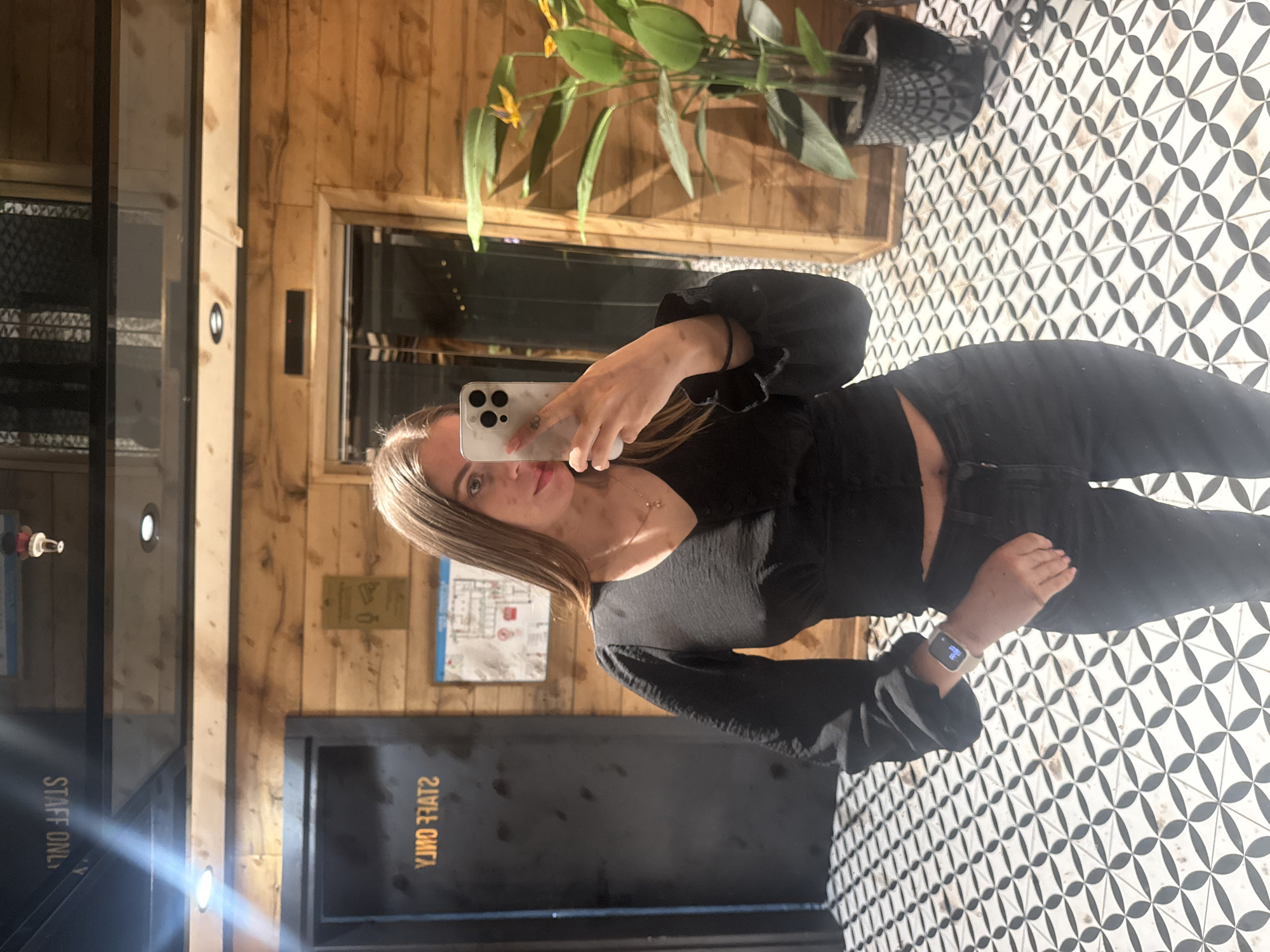 The image shows a woman taking a selfie in what appears to be a hallway or small room. She is holding a white phone in her right hand, partially obscuring her face. She is wearing a black long-sleeved top, black jeans, and a watch on her left wrist.
Behind her is a wooden paneled wall with a doorway on the left labeled "STAFF ONLY" in reverse. To the right of this doorway, there is a map or diagram affixed to the wall. A potted plant is situated in the upper portion of the frame. The floor is tiled with a geometric pattern of black and white. The location appears to be indoors, with the use of artificial lighting evident. The exact location in Batumi, Georgia, is not discernible from the visual information provided.