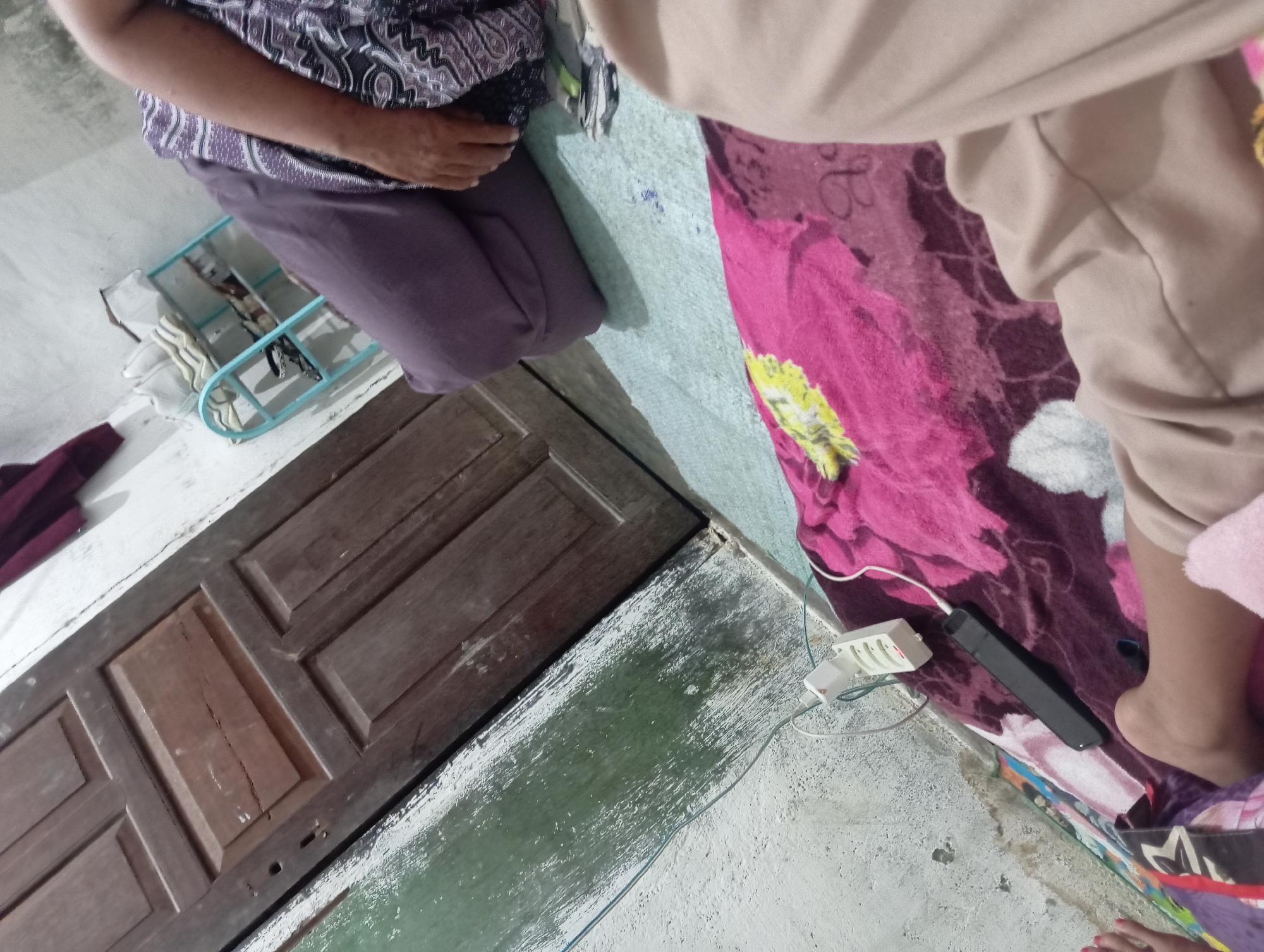 A person is lying on a patterned blanket on the floor of a room in Rejosari, Indonesia. A brown wooden door is visible behind the person, and a white electrical outlet strip with cables is plugged into the wall. There is a light blue shelf unit with items on it to the right of the door. The person is wearing a purple garment, and their legs and feet are visible. The overall impression is of a casual indoor setting.
