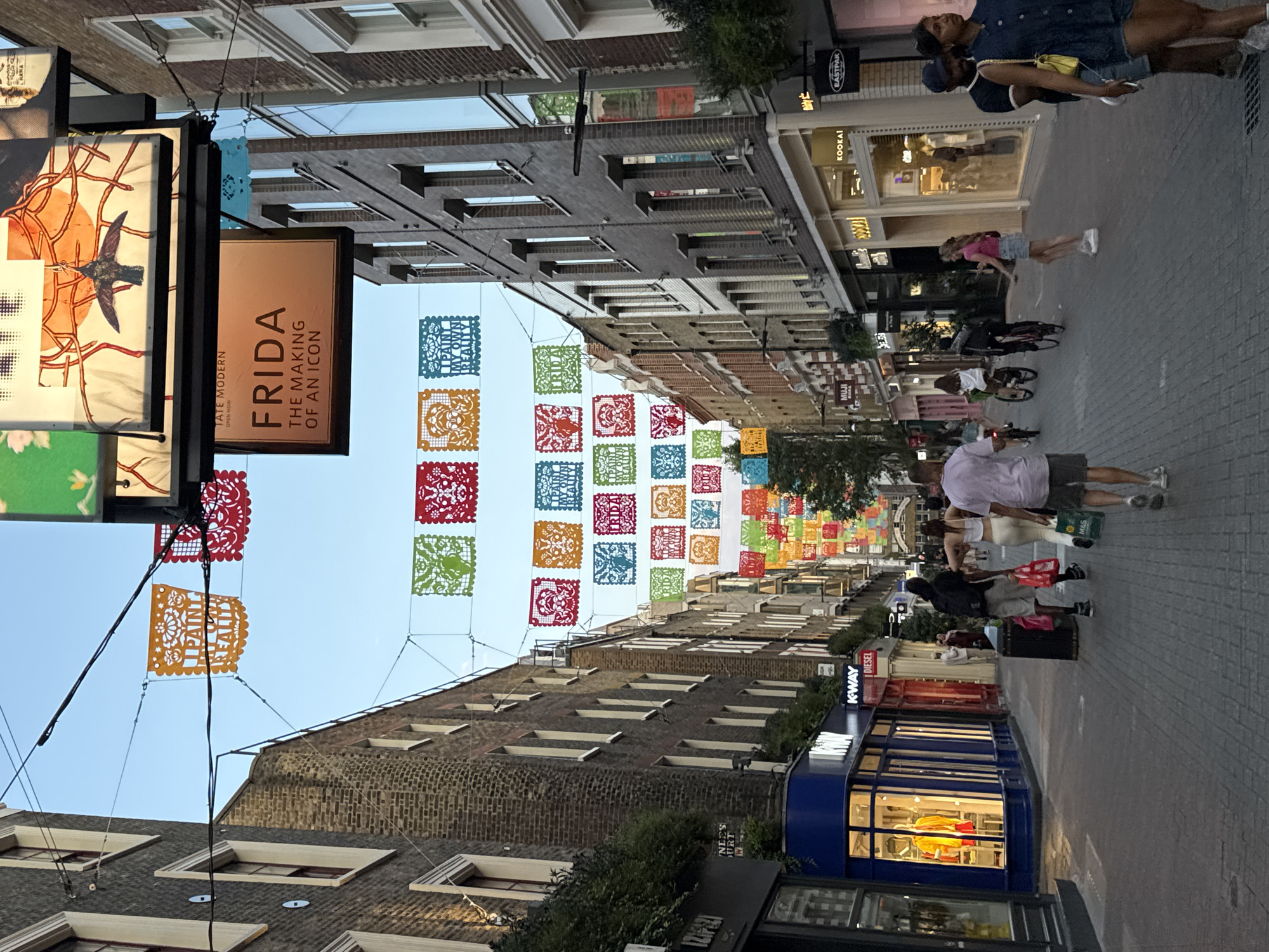 This is a vertical, eye-level, outdoor shot taken on a street in London, United Kingdom. The time of day appears to be late afternoon or early evening, as indicated by the soft, warm lighting and the lengthening shadows. The sky is clear and blue.
The scene is a bustling street lined with shops and buildings, adorned with colorful papel picado banners strung across the street. These banners, common in Mexican folk art, are intricately cut and feature various designs and patterns. They create a festive and vibrant atmosphere.
Several people are visible walking along the sidewalk and street. Some are casually strolling, while others appear to be shoppers or tourists. One person is riding a bicycle. The people are dressed in casual summer attire.
Prominently displayed on a sign is "FRIDA THE MAKING OF AN ICON" from TATE MODERN, suggesting an exhibition or event related to Frida Kahlo. Other visible text includes shop names like "KOOKAI," "MUJI," "EASTPAK," and "DIESEL," indicating a commercial area. The architecture of the buildings is varied, with some featuring traditional brickwork and others displaying more modern shopfronts with large windows.
The overall mood of the image is lively and dynamic, capturing the energy of urban life combined with a touch of cultural celebration. The composition draws the viewer's eye down the street, past the hanging decorations and shops, towards the distant buildings.