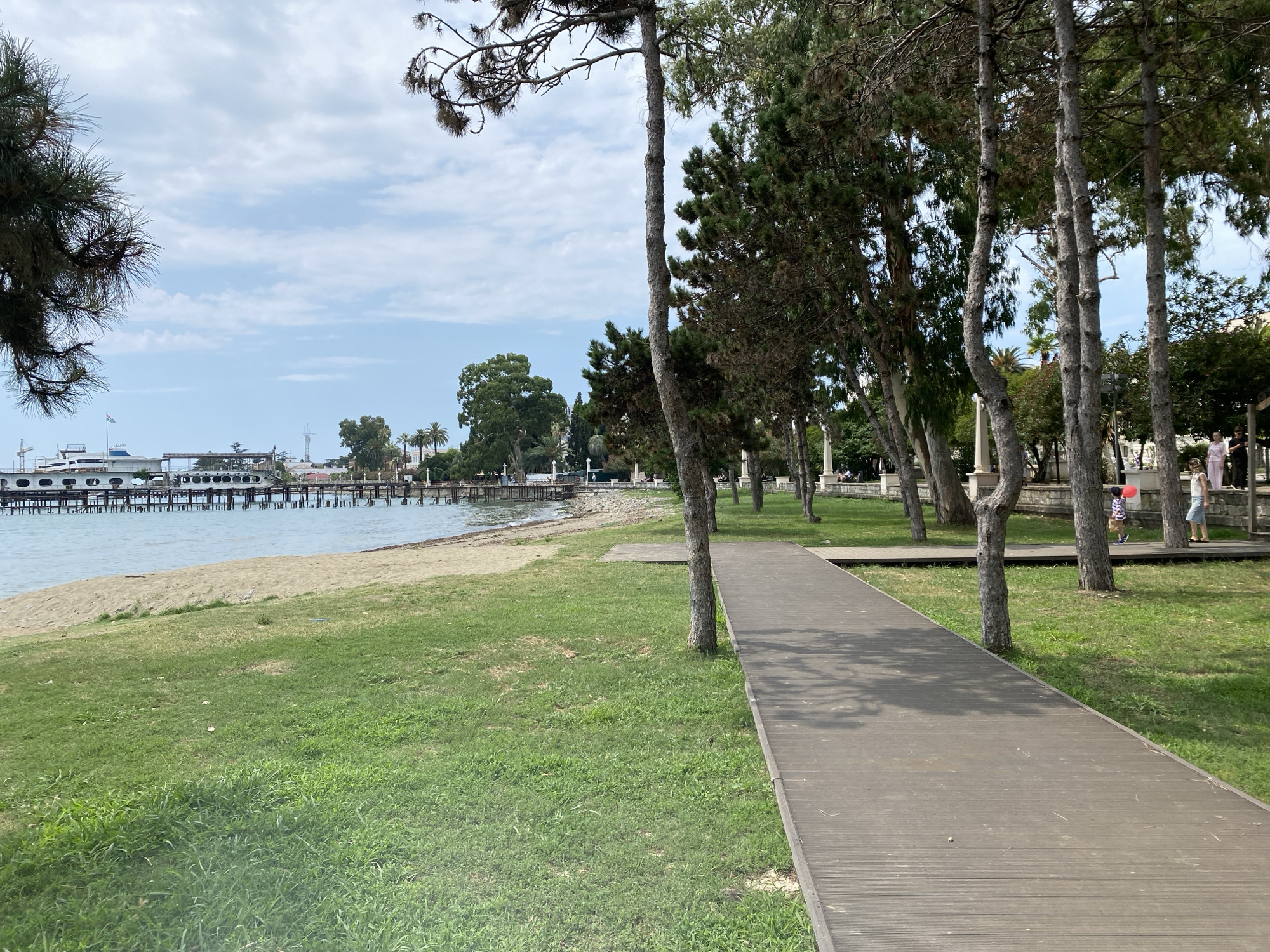 The image depicts a boardwalk path running alongside a grassy area and a beach. The boardwalk is constructed of dark wooden planks and curves slightly to the right. Several trees with slender trunks are positioned along the edge of the boardwalk, casting shadows on the path. To the left of the boardwalk is a strip of green grass, followed by a sandy beach and then the sea. In the distance, a long pier extends into the water, with a white boat docked at its end. The sky is partly cloudy with a pale blue hue. In the background, beyond the trees, are some buildings and palm trees, suggesting a park or promenade area. Further to the right on the boardwalk, two individuals are visible: a child holding a red balloon and an adult walking away from the camera. The scene appears to be a public space, possibly a tourist area in Sokhumi, Abkhazia, given the presence of the boardwalk, beach, and pier.