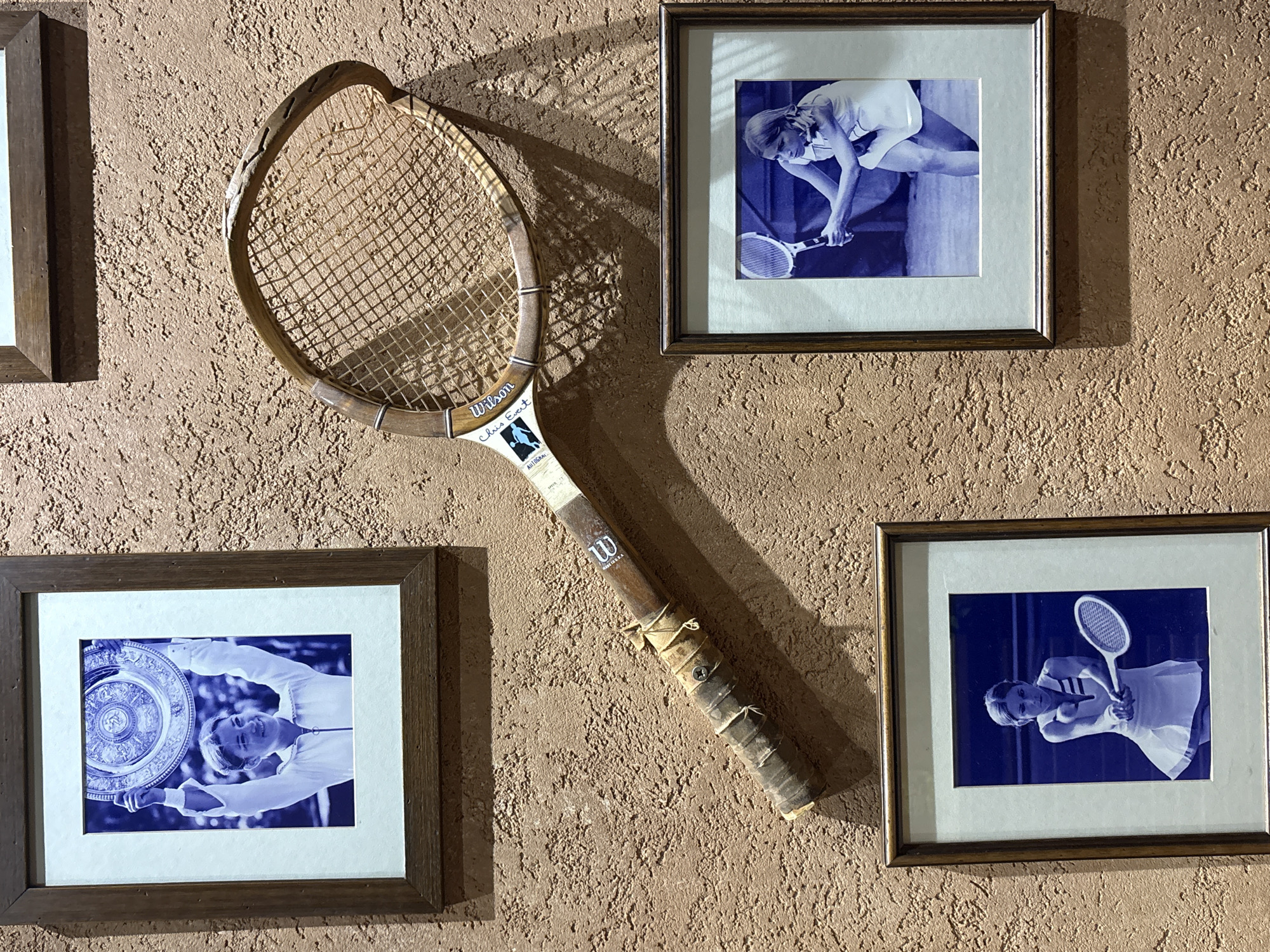 A wall display features a vintage wooden tennis racket and three framed, blue-toned monochrome photographs of female tennis players, all set against a textured, reddish-brown stucco wall. The specific location within Cukarica Urban Municipality, Serbia, cannot be determined from the image content.
The central object is an aged, light-colored wooden tennis racket, labeled "Wilson" and "Chris Evert" on its throat. The racket shows significant wear, including a damaged and cracked section on the upper right side of its head and a worn, fabric-wrapped grip secured by a visible screw and cord. It is mounted diagonally on the wall, extending from the lower-left to the upper-right quadrant.
Above the racket, a framed photograph depicts a smiling woman in a white tennis outfit, holding a large, ornate silver trophy (resembling the Wimbledon Venus Rosewater Dish) overhead with both hands. The background is blurred, suggesting an outdoor setting.
To the lower left of the racket, another framed photograph shows a woman in a white tennis dress with a focused expression, poised to execute a two-handed backhand shot.
To the lower right of the racket, a third framed photograph features a woman in a white tennis outfit in a low, athletic stance, holding her racket with both hands, appearing ready to strike the ball.
All three photographs are matted with a light blue border and enclosed in dark brown wooden frames. Distinct shadows are cast on the textured wall from the framed items and the tennis racket, indicating a light source from the upper-left.