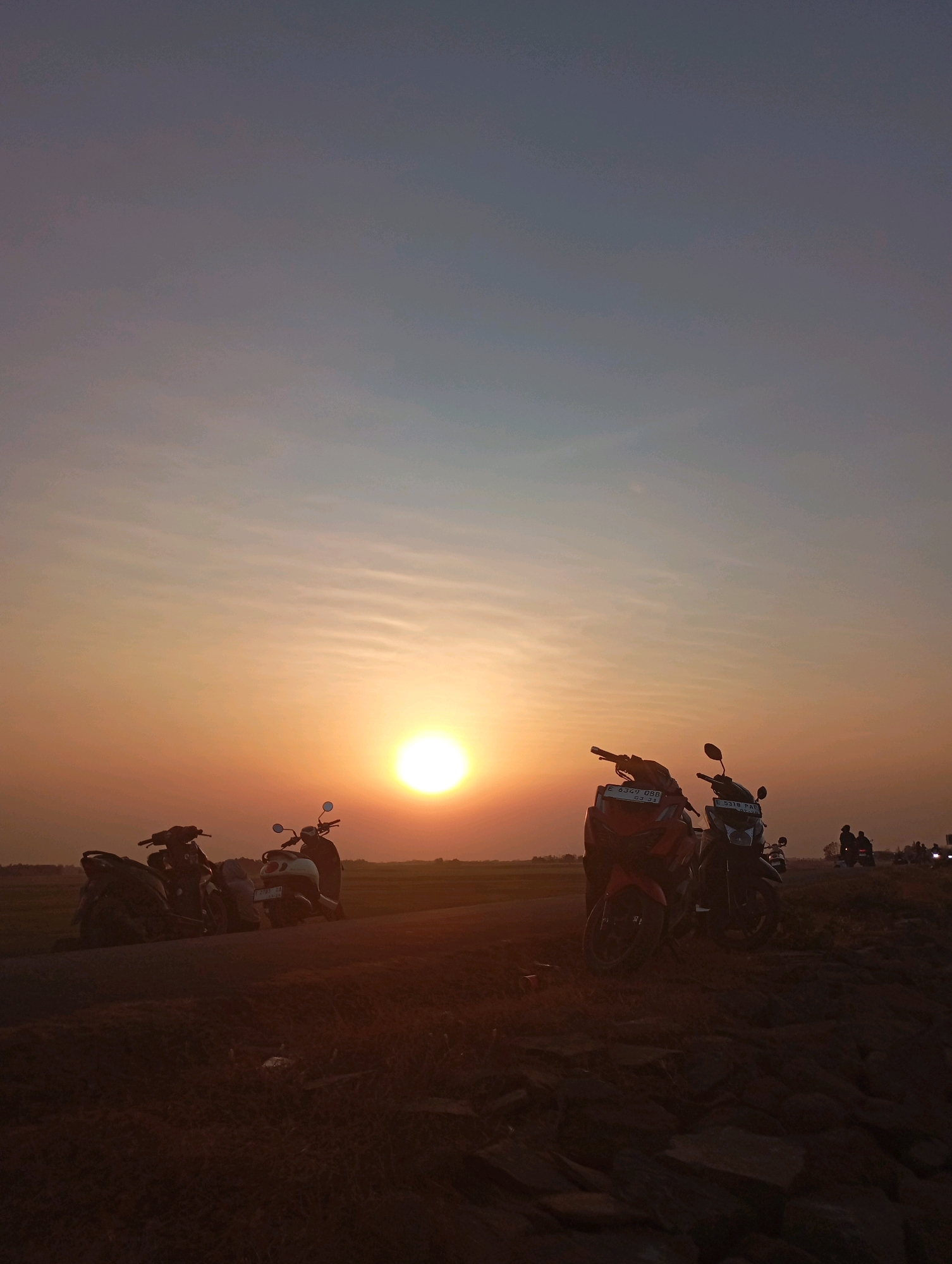 A vertical, eye-level shot captures a sunset over Haurkolot, Indonesia. The sun, a bright orb, is positioned in the middle left of the frame, casting a warm orange glow across a hazy sky with horizontal cloud streaks. In the foreground, several parked motorcycles are silhouetted against the sunset. Two prominent motorcycles are in the center, one red and the other dark. To the left, a group of darker motorcycles are clustered together. In the distance, on the right side of the image, several figures on motorcycles are visible, appearing as dark silhouettes against the horizon. The ground in the foreground is rough and uneven, with scattered rocks and dry vegetation. The background suggests a rural or semi-rural setting with flat land that transitions into darker silhouettes of distant features. The overall mood is tranquil and serene, characteristic of a sunset in an open landscape.
