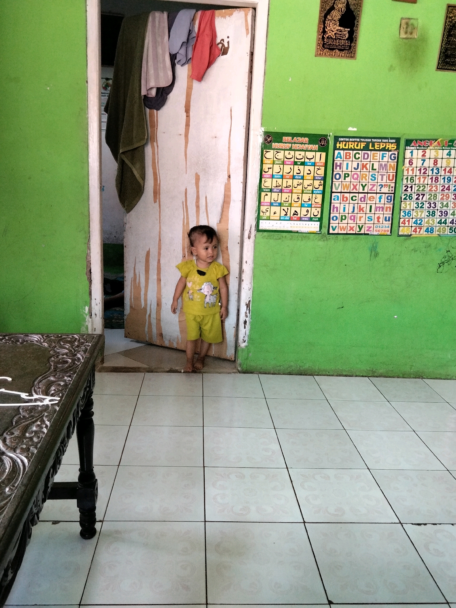 A young child, appearing to be around two to three years old, stands in a doorway. The child is wearing a yellow t-shirt with a graphic print and matching yellow shorts. They are barefoot and looking to the right. The doorway opens into a room with a darker interior. The child is positioned in front of a light-colored door with vertical wood grain and some visible damage. To the left of the doorway, a wall is painted bright green. On the right wall, three educational posters are displayed: one with Arabic script, one with the English alphabet in both upper and lower case, and one with numbers from 1 to 50. Above these posters, there are two framed religious artworks. In the foreground, a dark wooden table with ornate carvings is visible on the left. The floor is tiled with a light-colored pattern. The scene appears to be indoors, possibly in a home or educational setting, in Rahong, Indonesia.