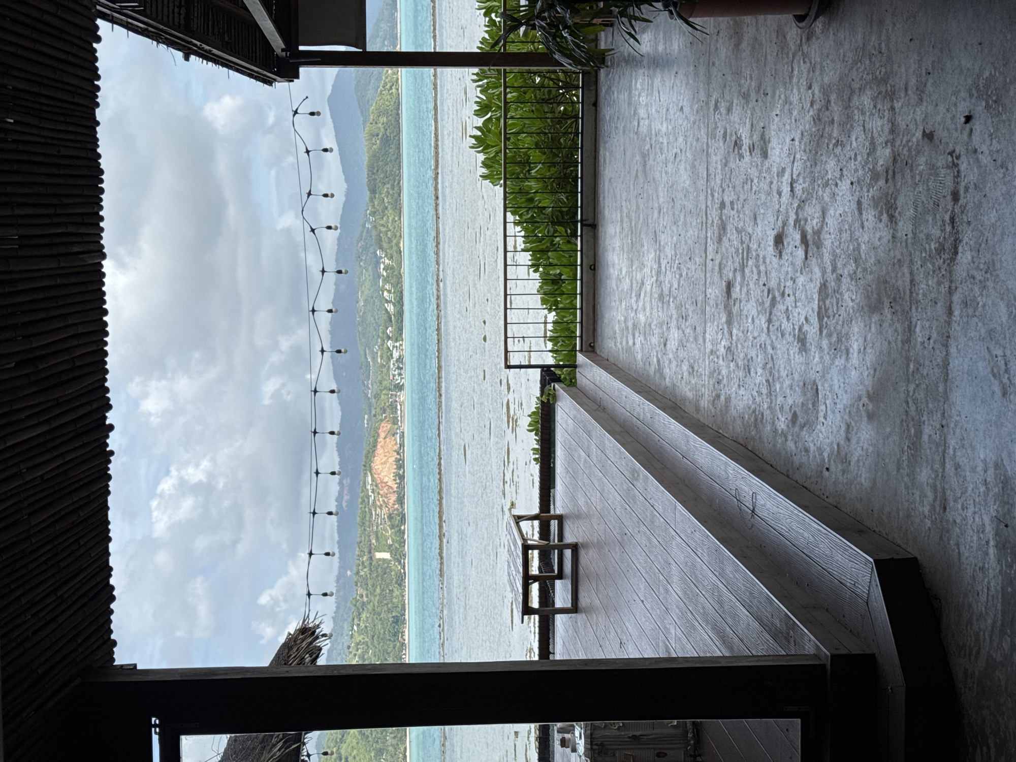 The image depicts a view from a covered outdoor area overlooking a body of water and a hilly, tree-covered landscape. In the foreground, a wooden deck with tiered steps leads down to a concrete floor. A simple wooden table sits on the deck. A black metal railing with green foliage behind it separates the deck area from the water. String lights are suspended overhead. The water is a vibrant turquoise color, with a lighter, possibly shallow area closer to the shore. In the distance, rolling hills covered in lush vegetation rise up, with some buildings visible on their slopes. The sky is overcast with dramatic clouds. The scene suggests a tropical or coastal resort setting.