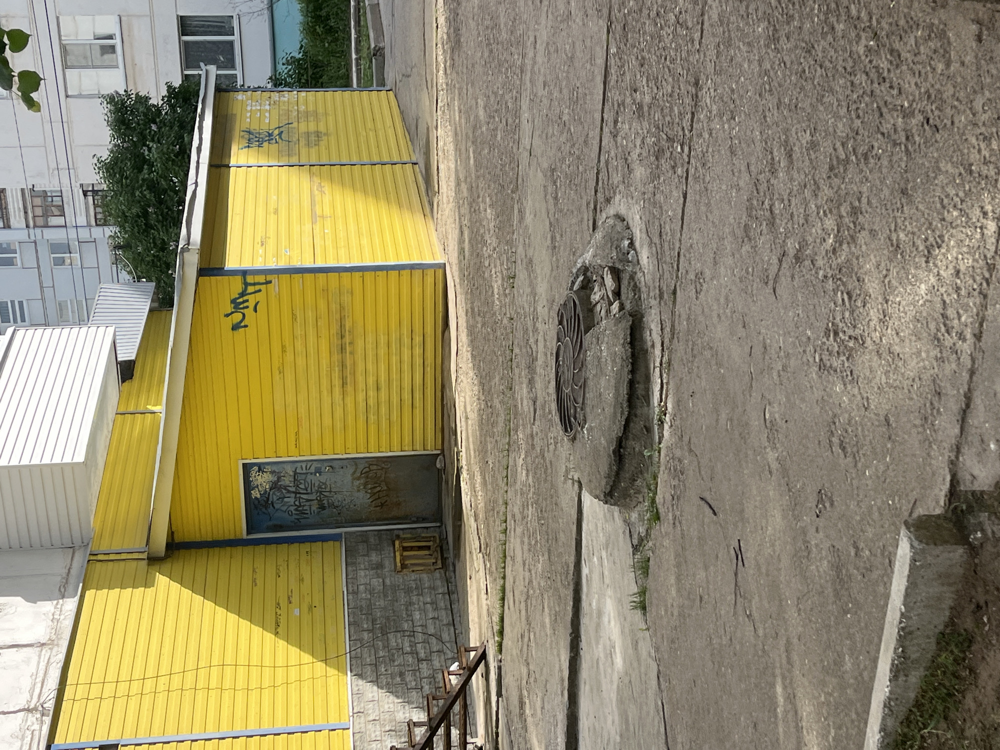 The image shows a section of urban landscape, likely in the city of Chaikovsky, Russia. A bright yellow building with corrugated metal siding dominates the left side of the frame. Graffiti is visible on the building's walls. The building has a blue-framed door and a possible window structure below it. To the right of the building, a cracked concrete sidewalk stretches towards the top of the image. Set into the sidewalk is a partially destroyed manhole cover. The overall atmosphere suggests a somewhat neglected or aged urban environment. Part of another building is visible in the background.