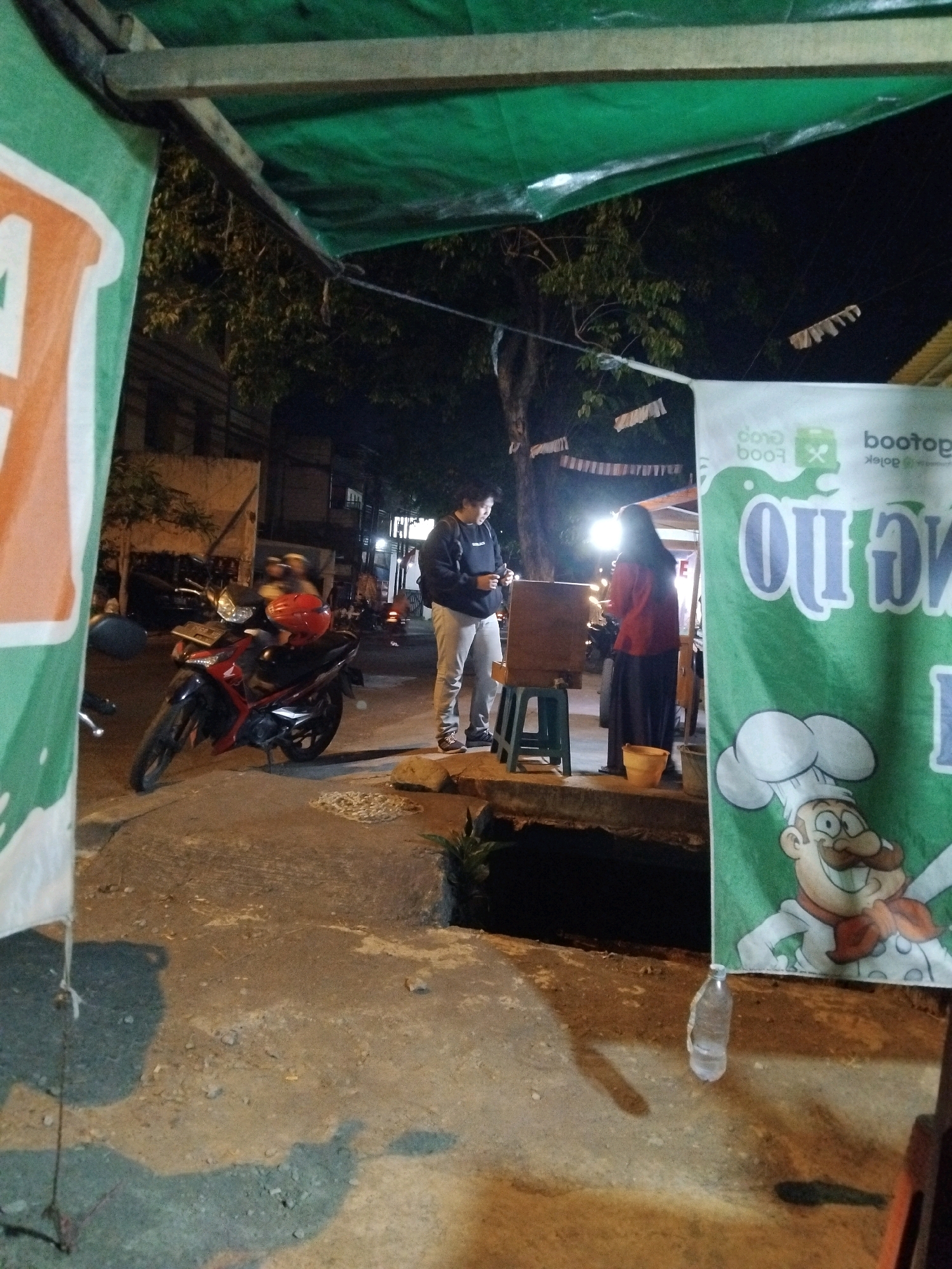 In Wedoro, Indonesia, at night, a person stands on a paved sidewalk beside a parked motorcycle. The motorcycle is red and black with a red helmet resting on its seat. The person is wearing a black jacket and light-colored pants, and appears to be looking at a mobile phone. Behind them, a food stall is set up with a green tarp overhead supported by wooden beams. A banner with a cartoon chef illustration and text is displayed on the right side of the stall. Another person, wearing a red top and dark skirt, is visible near the stall. The area is lit by artificial lights, casting shadows on the ground. In the foreground, a plastic water bottle sits on the pavement. Trees and buildings are visible in the background.