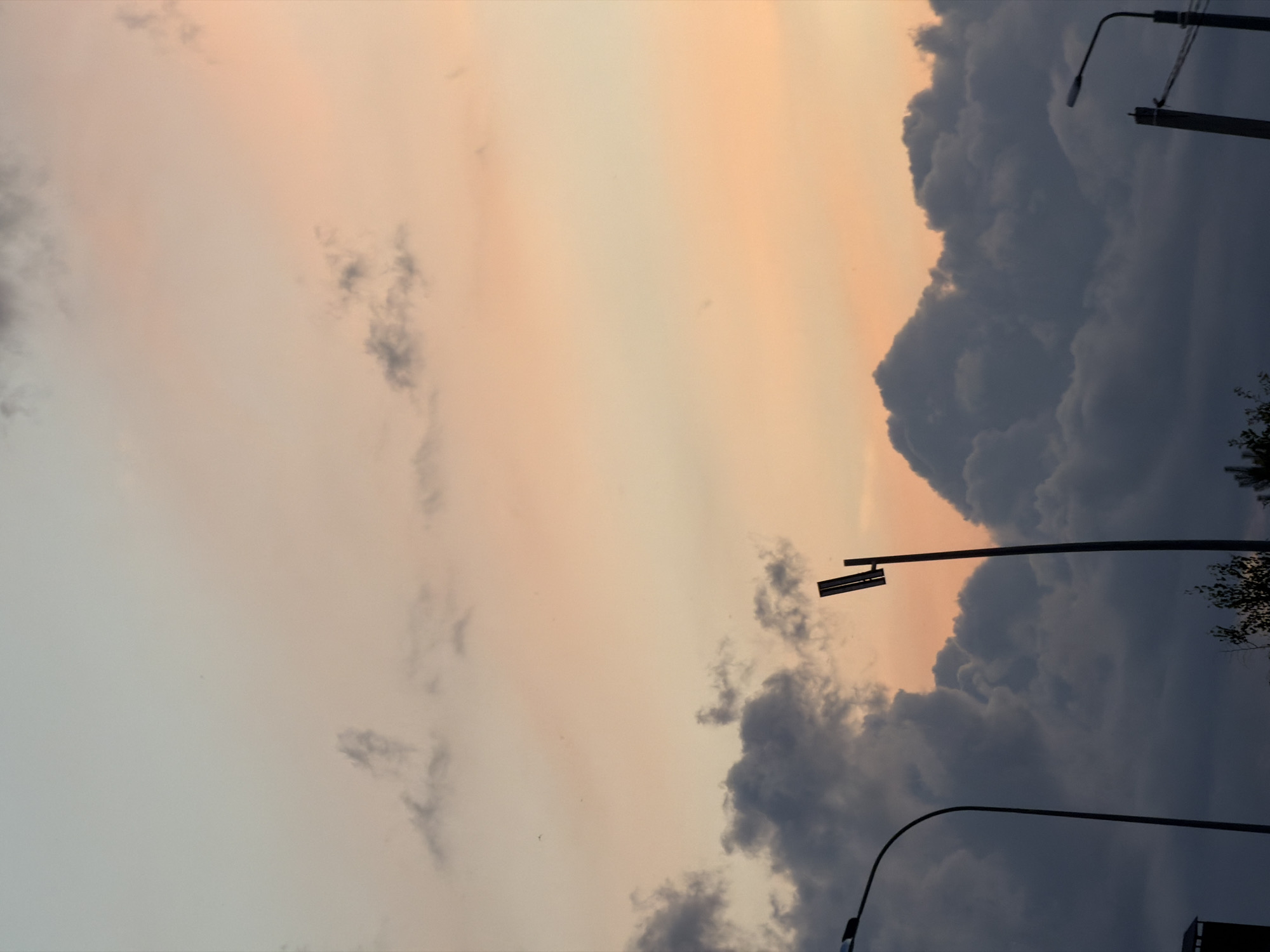 The image captures a dusky sky with scattered clouds, tinged with hues of pale blue and soft peach, suggesting either dawn or dusk. Two streetlights are visible in silhouette against the sky. The streetlight on the left is a curved arm extending from a pole, while the one on the right is a straighter pole with a rectangular fixture. Dark, dense clouds occupy the lower portion of the frame, contrasting with the lighter sky above. Hints of greenery or treetops can be seen at the very bottom edge of the image. The scene appears to be an outdoor urban or suburban environment in Laishevo, Russia.