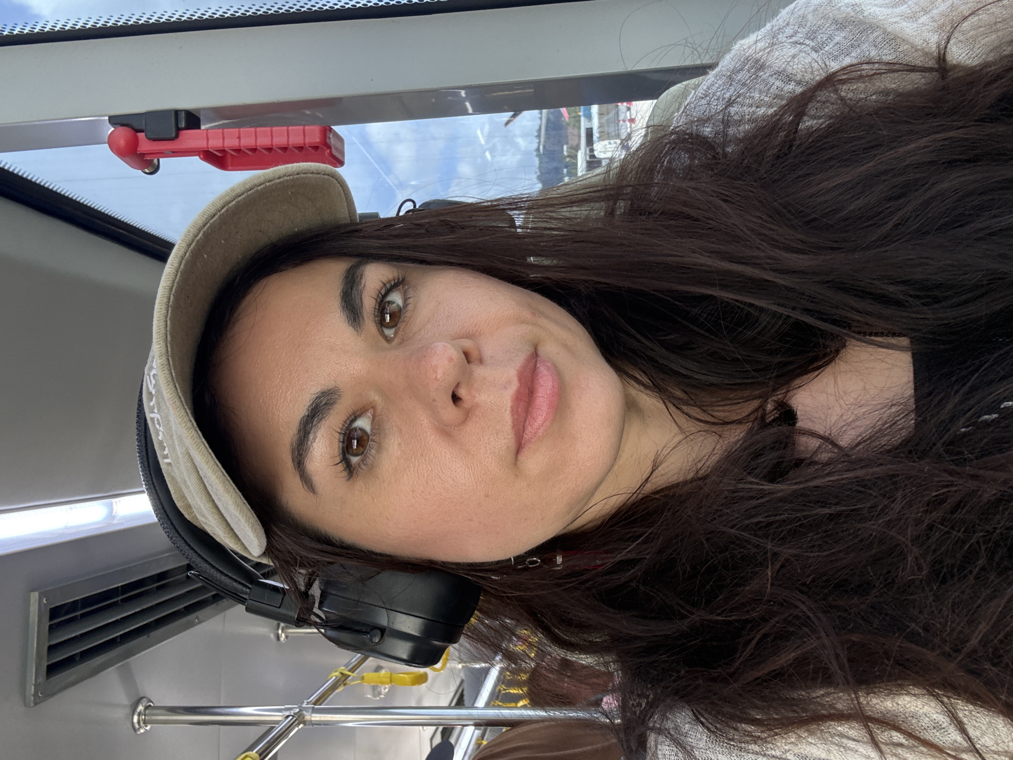 In a close-up, selfie-style photograph, a woman with long, dark, wavy hair is depicted from the chest up. She is wearing a light-colored baseball cap tilted forward, obscuring some of her forehead. Over her ears, she has black headphones on. She is looking directly at the camera with a neutral expression, her eyes are brown and her lips are slightly parted. She is wearing a light-colored, textured cardigan over a black top.

The background suggests an indoor setting with hints of an outdoor view through a window. Above and behind her head, metal bars and yellow straps are visible, possibly part of public transportation seating. To the right of her head, a red emergency hammer is mounted on a white pole next to the window. Through the window, a cloudy sky and a distant landscape with buildings and what appears to be a harbor or waterfront can be seen. The lighting in the scene is bright, indicating daylight.

The image does not provide any specific visual cues that can definitively place the scene in Sakhu, Thailand.