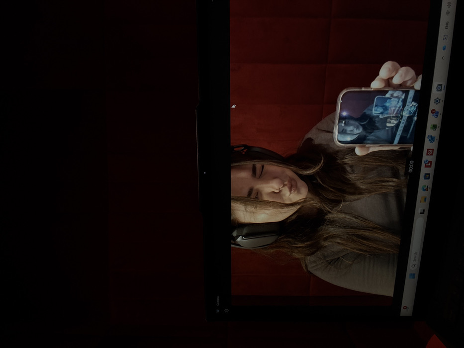 The image displays a close-up of a laptop screen, which is actively showing a live webcam feed of a woman. This scene is located in Bnei Brak, Israel.
**Laptop Interface:** The top bezel of the black laptop screen displays the text "Camera" and a small light indicator. The bottom portion of the screen shows part of the Windows taskbar, including a search bar, several application icons (e.g., Microsoft Edge, settings, photos), and a digital timestamp "00:00". A white mouse cursor is visible near the top right of the webcam feed area.
**Webcam Feed Content:** The main subject of the webcam feed is a young adult female with long, dark brown hair. She is wearing dark, over-ear headphones and a grey, long-sleeved shirt. Her facial expression is a puckered or "pouty" pose, with her eyes partially squinted. She is holding a smartphone in her left hand (viewer's right), extended towards the camera. The smartphone has a reflective, light-colored case. Its screen clearly displays a reflection of the woman holding it, with a smaller, recursive reflection of the phone itself visible within that reflection, demonstrating a Droste effect.
**Background (within webcam feed):** Behind the woman is a wall covered in dark red, rectangular panels, arranged in a grid pattern.
**Environment:** The ambient lighting around the laptop is dim, contrasting with the illuminated scene displayed on the screen.