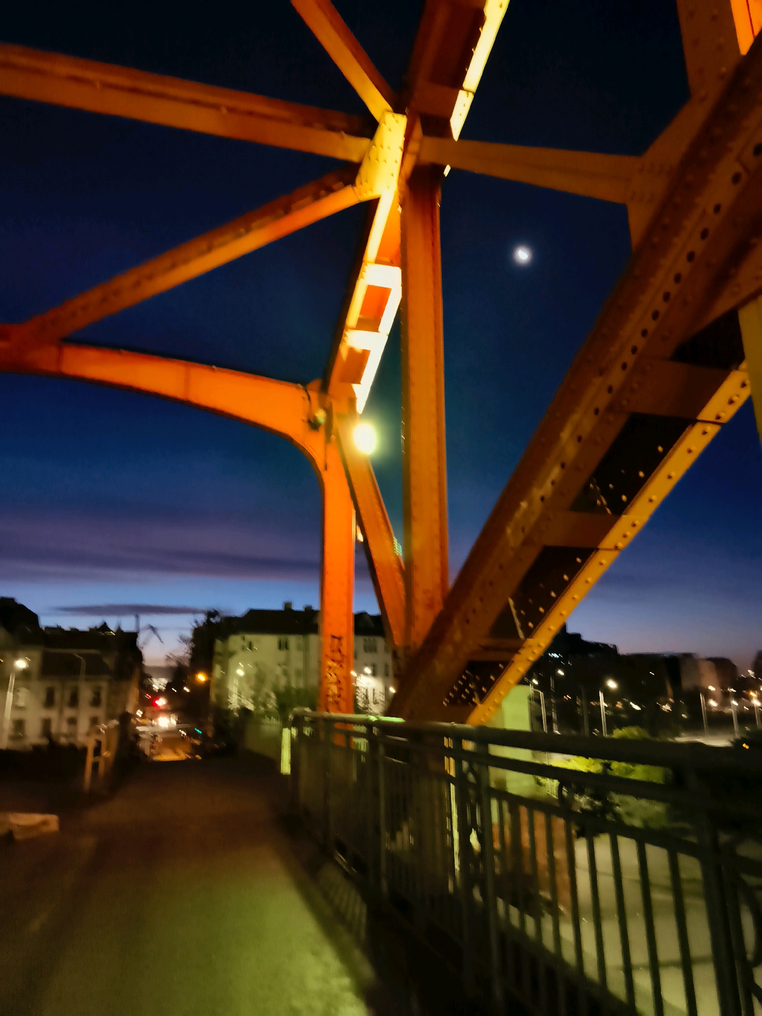 The image captures a nighttime view from a pedestrian walkway on a bridge in Gdańsk, Poland. The bridge's bright orange, angular metal structure dominates the foreground, with its beams crisscrossing against a deep blue twilight sky. A crescent moon is visible in the upper right quadrant of the sky. In the background, streetlights illuminate buildings and a road with some traffic lights visible. The walkway itself is dimly lit, with yellow and green hues cast by unseen light sources, reflecting off the wet surface. A metal railing lines the right side of the walkway. The overall impression is one of an urban landscape transitioning from day to night.