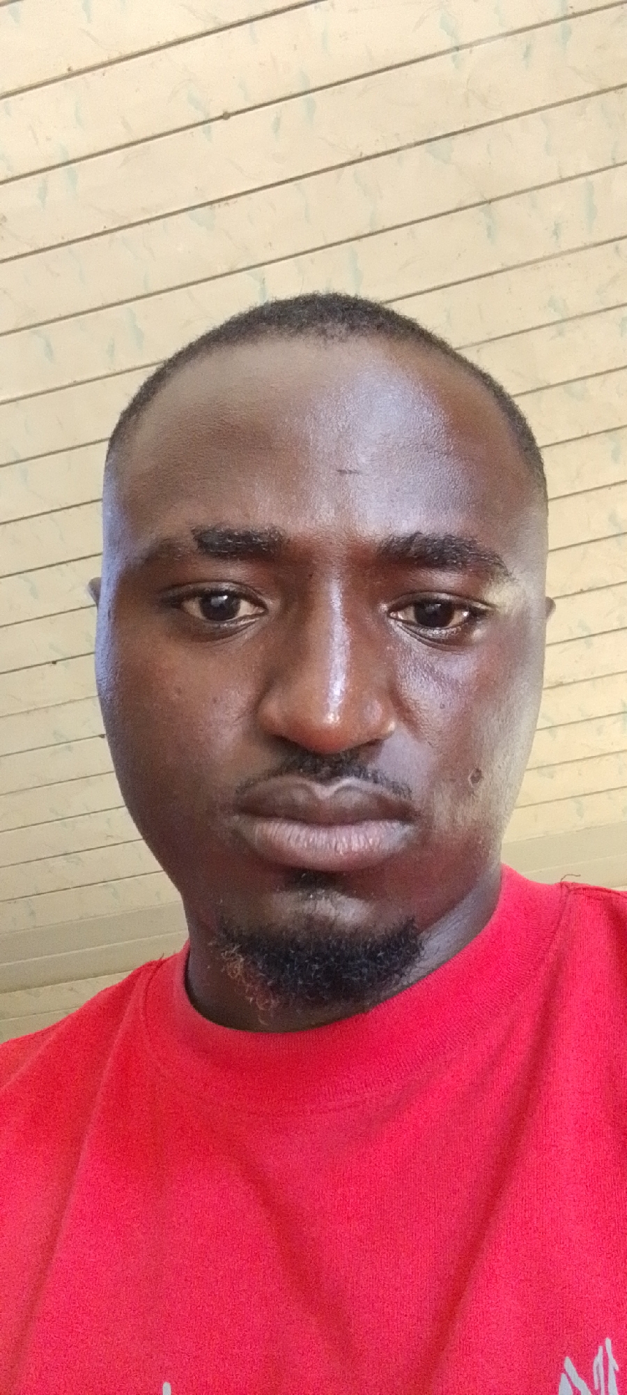 A close-up shot of a dark-skinned man with short hair and a trimmed goatee. He is wearing a red t-shirt. The background is a light-colored ceiling with visible lines. The photograph appears to be taken indoors, possibly in Vom, Nigeria, as that location was specified in the prompt.
