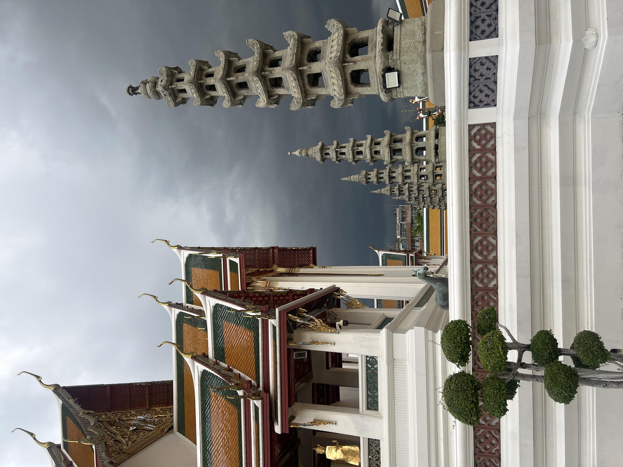 This is a ground-level, medium shot of Wat Arun in Bangkok, Thailand. The temple's ornate, multi-tiered roofs with vibrant orange and green tiles are prominent on the left side of the frame. The structure features white walls and decorative gold accents. In the foreground, a sculpted bonsai tree stands to the left, with its branches shaped into rounded forms. Behind it, a white balustrade with intricate red and grey decorative panels separates the foreground from the middle ground.
In the middle ground, a bronze sculpture of a horse is visible, facing left. Further back and to the right, a series of tall, weathered stone stupas of varying heights rise against a dark, cloudy sky. The largest stupa is on the far right, and a line of progressively smaller stupas recedes into the distance. These stupas are intricately carved with multiple tiers and openings. In the far background, hints of other temple structures with orange roofs and urban buildings can be seen. The sky is overcast and dark, suggesting an impending storm. There are no people visible in the image. The overall scene depicts the architectural grandeur of the temple complex under a dramatic sky.