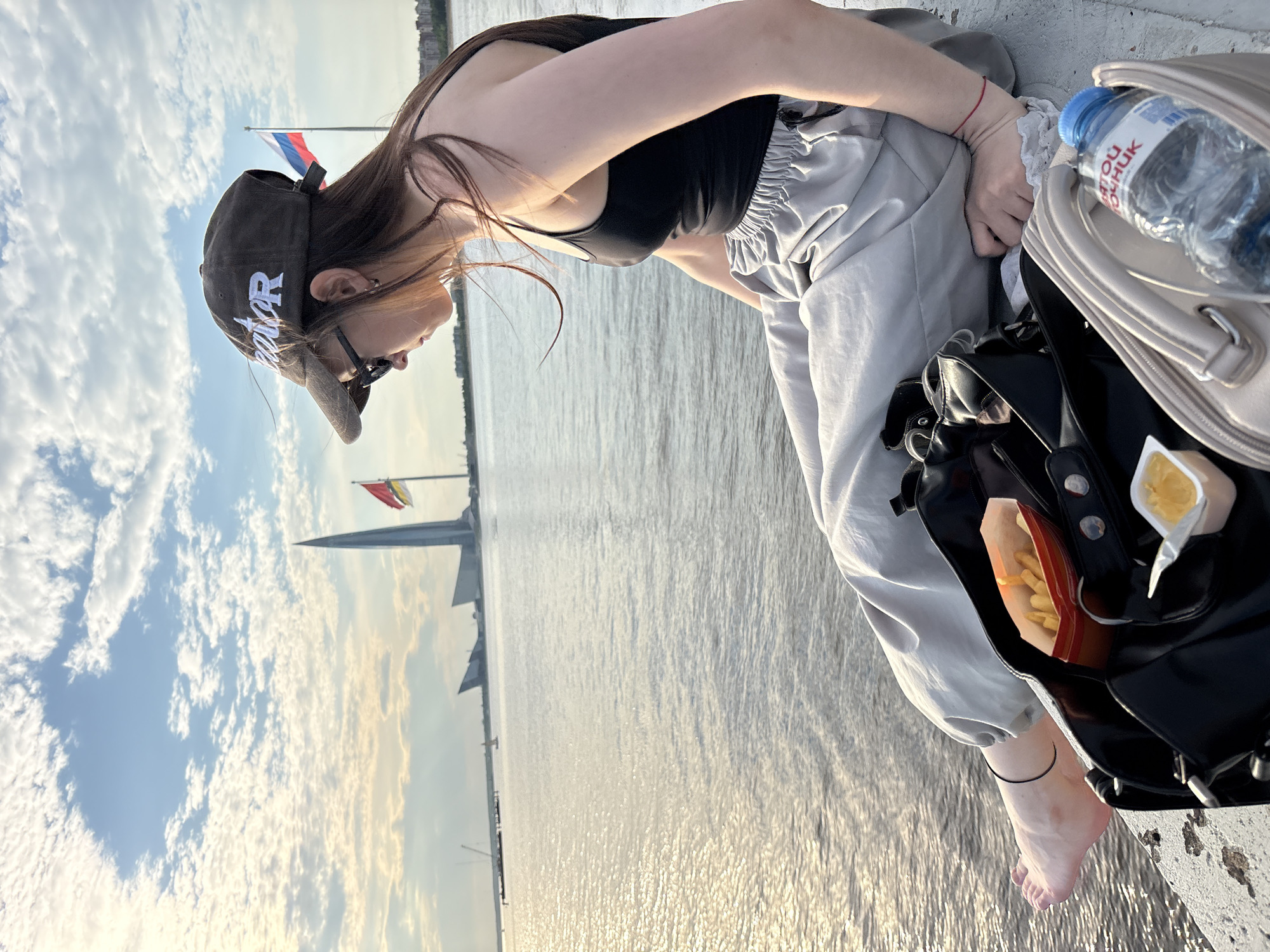 A young woman is captured leaning forward outdoors, facing away from the camera's direct view. She has long dark hair, a black tank top, and light grey baggy pants, with one bare foot visible, adorned with a thin black anklet. She wears glasses and a dark baseball cap with the word "Creator" in white script. On her wrist, a thin red bracelet is visible.
She is positioned on a concrete edge overlooking a wide body of water that glistens with sunlight, indicating a bright daytime setting, possibly late afternoon. Beside her, a black bag is open, revealing a plastic bottle of "СВЯТОЙ ИСТОЧНИК" (Svyatoy Istochnik) branded water, a paper container of french fries, and a small clear cup of condiment.
In the background, a blue sky with scattered white clouds stretches above. Two flags fly from poles: one is the Russian flag (white, blue, red horizontal stripes), and the other has horizontal stripes of yellow, red, and yellow. A distinctive, tall modern skyscraper, likely the Lakhta Center, is visible in the distance across the water. The scene portrays a calm moment by the water, possibly on a pier or embankment in Saint Petersburg, Russia, with the woman appearing to be focused on her bag or enjoying a snack.
