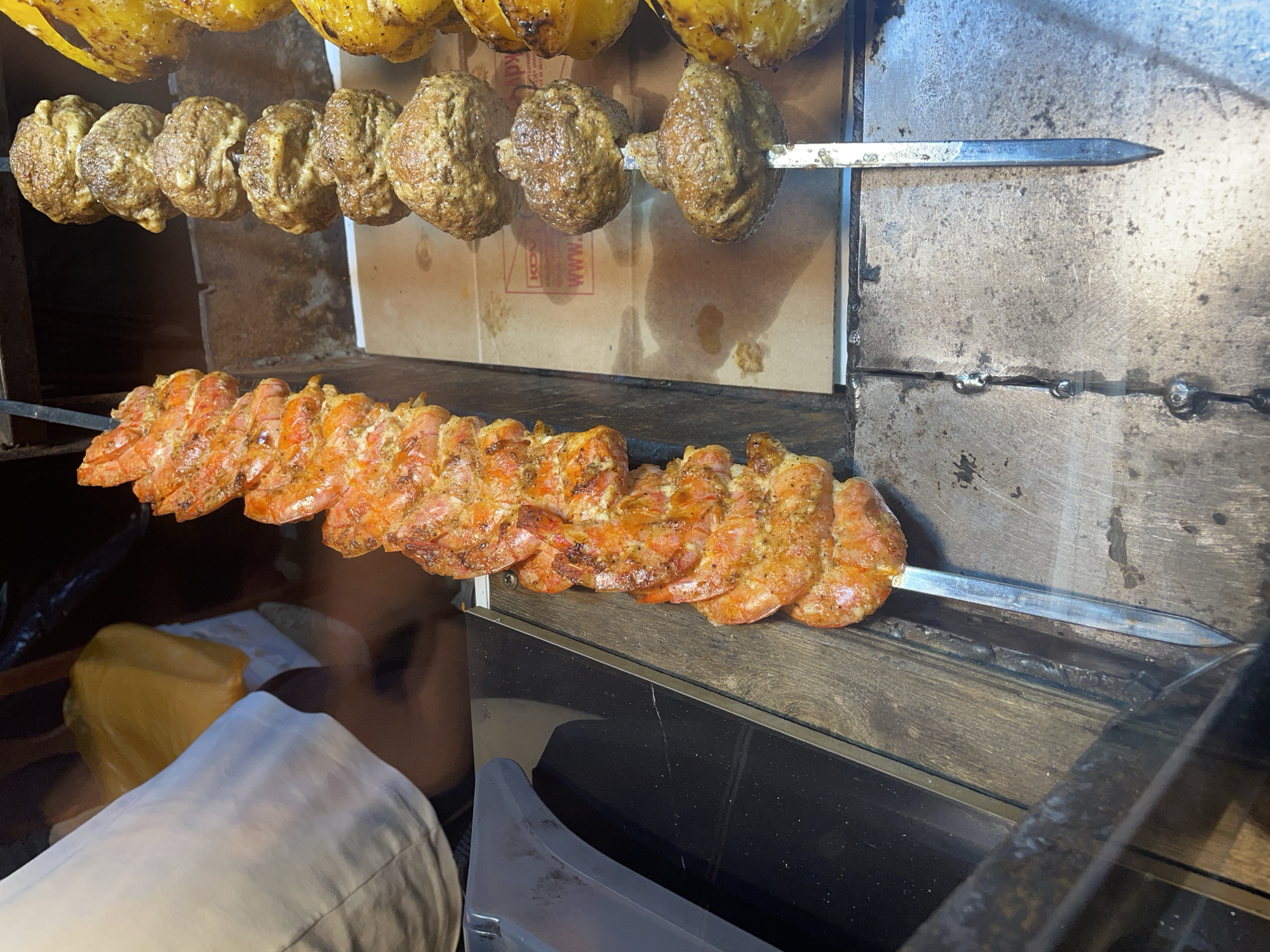 In the city of Unknown, Unknown, an overhead shot captures a close-up view of several skewers of cooked food. On the left, a skewer is densely packed with vibrant orange shrimp, each piece seasoned and appearing grilled. To its right, two more skewers are visible. One contains a row of round, brown, meatball-like items, also appearing to be grilled. The farthest skewer to the right holds what appear to be yellow bell peppers or a similar vegetable, also cooked. The food is displayed in what looks like an indoor food stall or market. The background includes a person's arm and torso wearing a white shirt, partially obscuring a yellow plastic bag. The food is arranged on metal skewers, with the bottom portions of two skewers visible resting on a metallic surface. The environment suggests a place where food is prepared and sold, with an emphasis on grilled items.