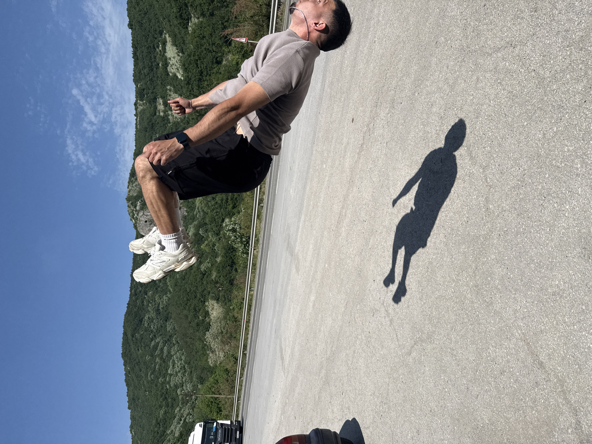 The image shows a man in mid-air, seemingly jumping or leaping. He wears a light brown short-sleeved shirt, black shorts, white socks, and white sneakers. A watch is visible on his left wrist. His shadow is cast on the ground to his right. The background consists of trees and a blue sky, with part of what appears to be a bridge or elevated road. A portion of a white truck or car is visible at the bottom edge of the frame. Given the prompt's information, the location is likely near the city of Targovishte, Bulgaria.