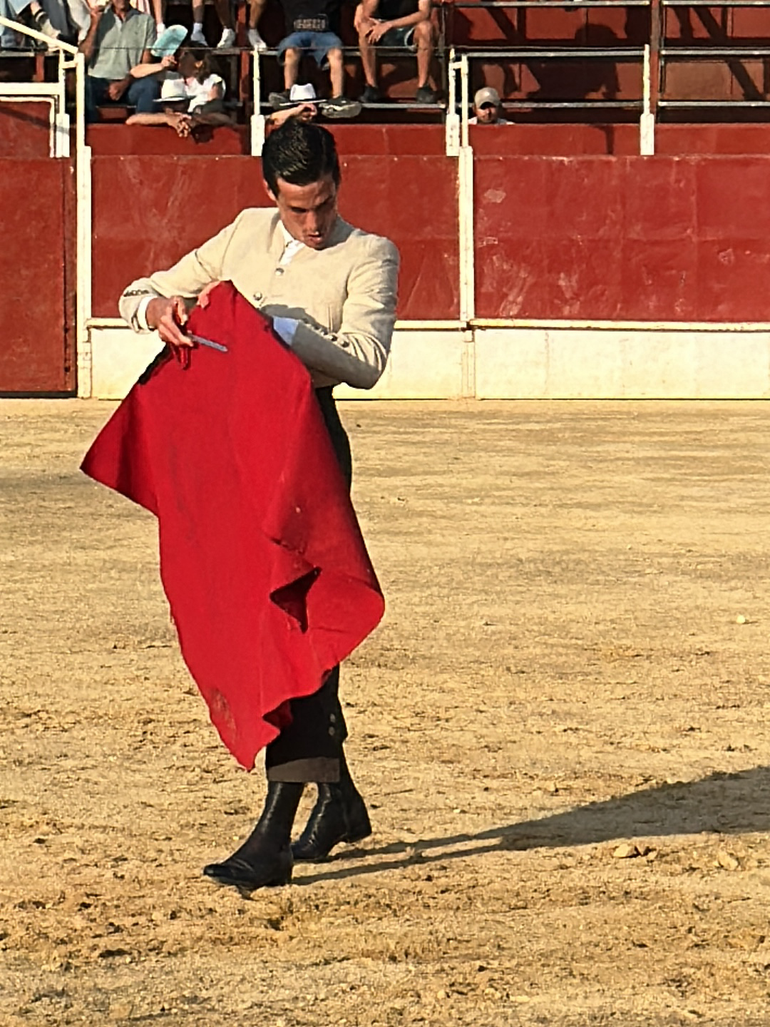 The image depicts a man in a bullfighting ring in San José del Valle, Spain. He is wearing a light-colored jacket, dark pants, and black boots, holding a red cape. The arena is a sandy surface with a high red wall surrounding it, and there are spectators visible in the background. The man appears to be adjusting the cape.