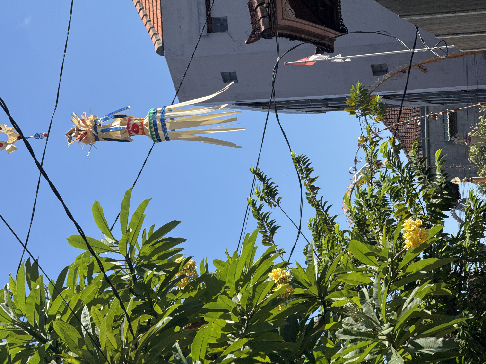In Kuta, Indonesia, a tall, decorative Balinese umbrella hangs suspended by wires in front of a grey building with a tiled roof. The umbrella, adorned with blue and white ribbons, features a conical top with gold and red accents, and long, pale yellow streamers cascading downwards. Green foliage, likely from a frangipani tree with yellow blossoms, partially obscures the view on the left. Electrical wires crisscross the bright blue sky. A small Indonesian flag, red and white, is visible on a pole to the right of the building, near a traditional Balinese architectural detail. The scene is illuminated by clear daylight, suggesting a warm, sunny day.
