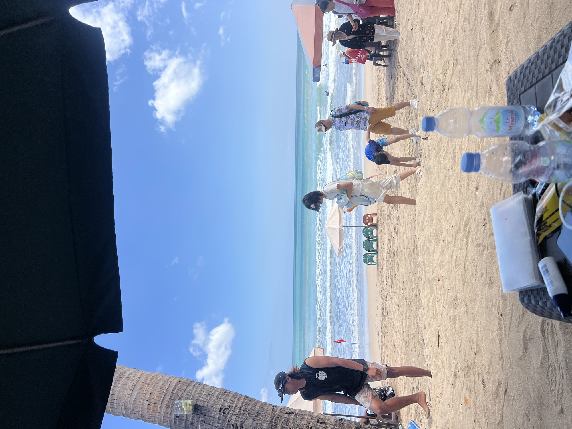 The image depicts a sunny day at Kuta Beach, Indonesia, with several people enjoying the sand and ocean. In the foreground, a table with two bottles of water, a packet of tissues, and other items is visible, suggesting a relaxed setting. A man wearing a black sleeveless shirt and patterned shorts stands near a palm tree to the left, looking out at the sea. Further down the beach, a group of people are walking, including a woman carrying a bag and a boy bending down. In the background, beach umbrellas and chairs are set up, and people are wading in the turquoise ocean, with waves rolling in. The sky is bright blue with scattered white clouds, characteristic of a tropical climate. The overall scene conveys a lively and pleasant atmosphere at a popular beach destination.