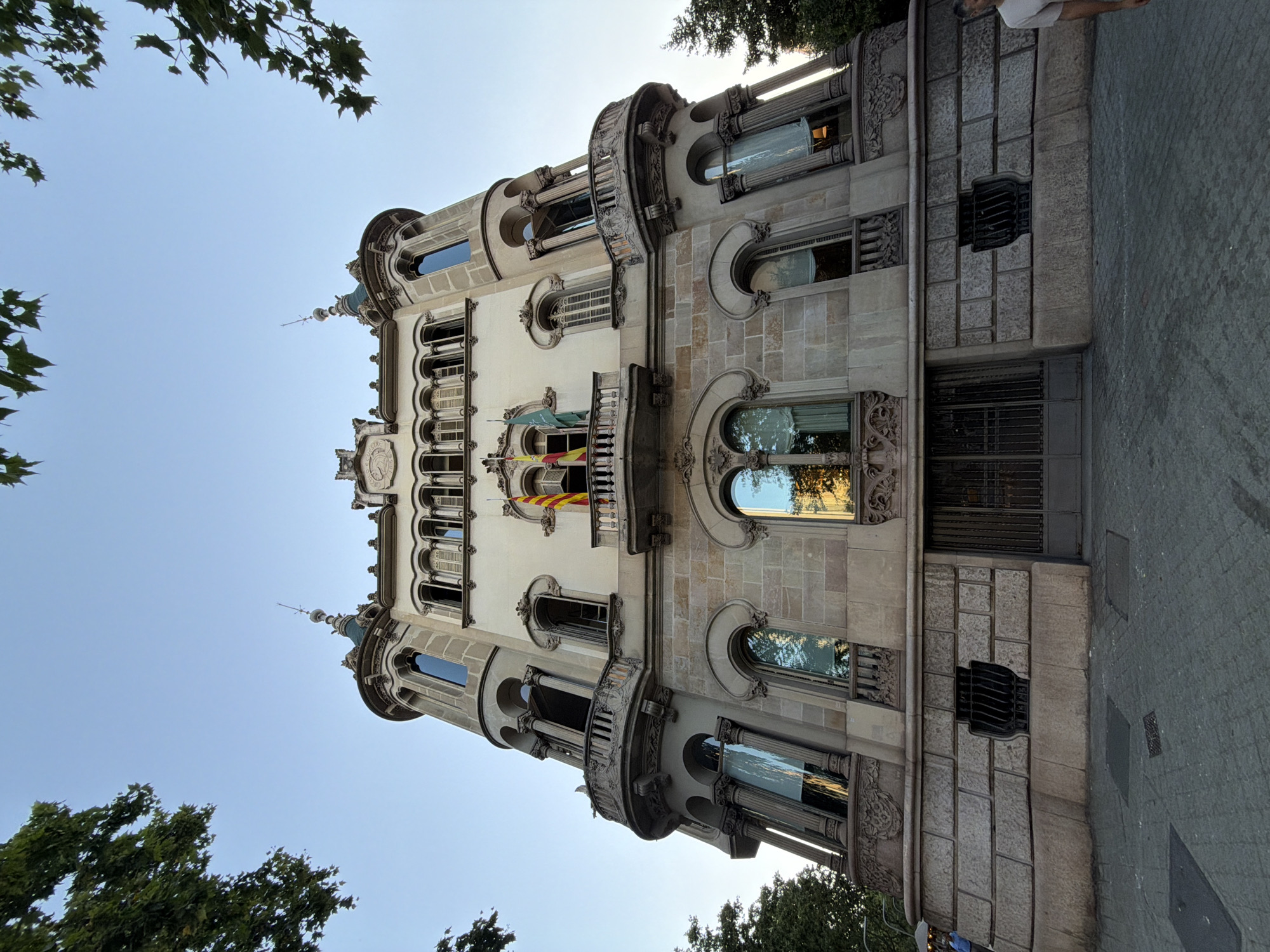 The image shows a low-angle, wide shot of the Casa de les Punxes, a modernist building located on Avinguda Diagonal in Barcelona, Spain. The building, constructed of stone and white stucco, features multiple towers with conical roofs, ornate balconies, and arched windows. Two flags, one yellow and red and the other green, are visible on a balcony. The sky is clear and blue, and trees with green leaves frame the top of the building. The street in front of the building is paved with cobblestones. A man in a light-colored t-shirt is standing on the right side of the image, looking towards the building.