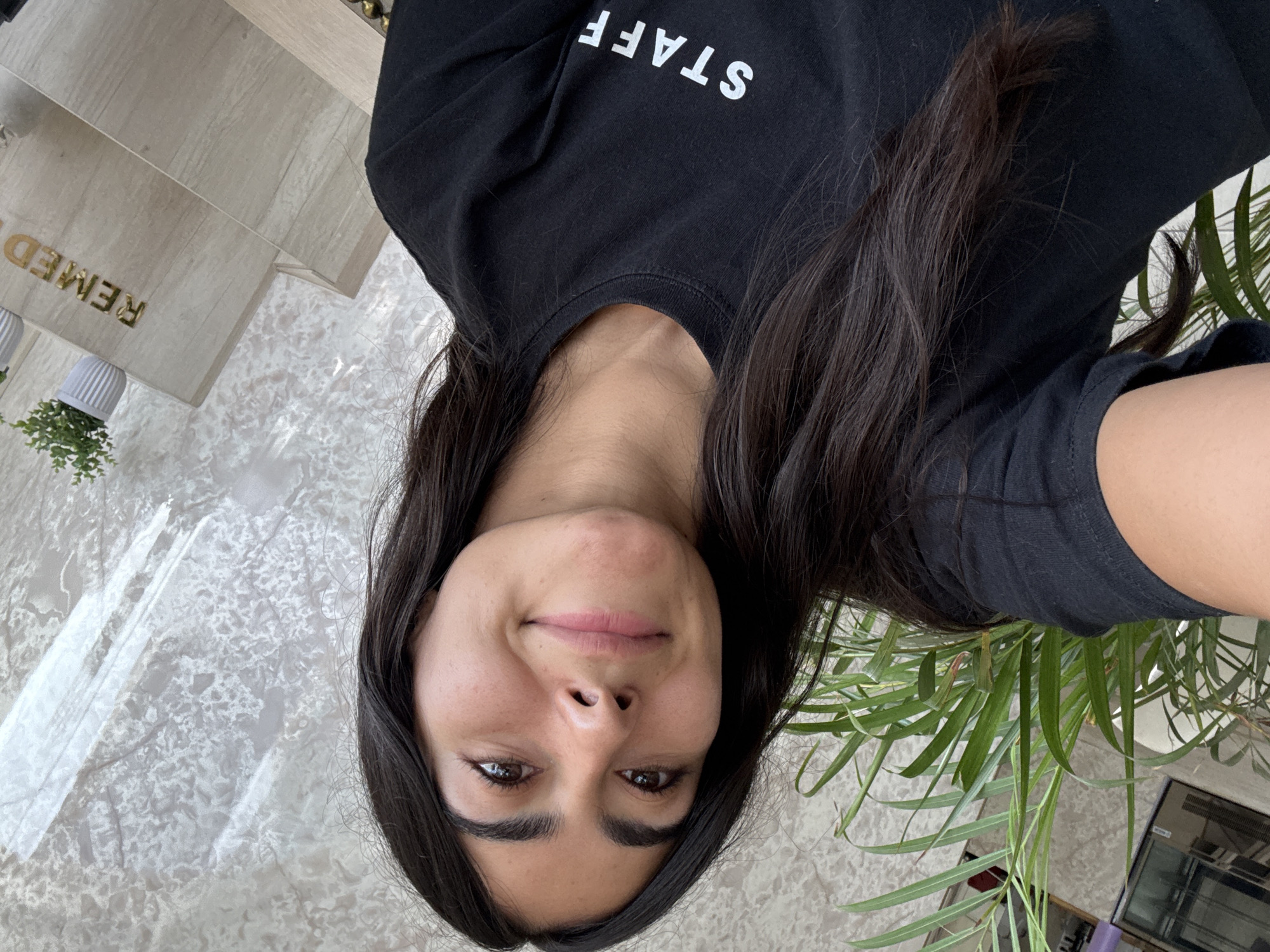 An eye-level selfie shows a young adult woman with long, dark hair, dark eyes, and a light complexion, looking up towards the camera with a subtle smile. She is wearing a black t-shirt with the word "STAFF" printed in white, capital letters across the chest.
The setting appears to be an indoor or semi-enclosed commercial space. To the woman's right (from her perspective, left of the frame when oriented), a large green plant with long, narrow leaves partially frames the shot. To her left, behind her, a section of a light-colored, possibly marble or tiled floor/wall with a mottled pattern is visible. Further back and slightly to the left, a light-colored wooden-textured surface features golden, embossed letters that spell "REMED" (likely part of a larger word like "REMEDY" or "REMEDIOS"). A small white, fluted planter with a small green plant sits near this surface. In the bottom right corner (when correctly oriented), a partial view of what seems to be a reflective surface or appliance, possibly a display or air conditioning unit, is visible.
The overall lighting suggests daytime. The woman's expression is calm and pleasant. The text "STAFF" on her shirt and "REMED" in the background hint at a professional or business environment, possibly a clinic or office in Puebla, Mexico. The activity is a self-portrait, captured from a low angle looking upwards.