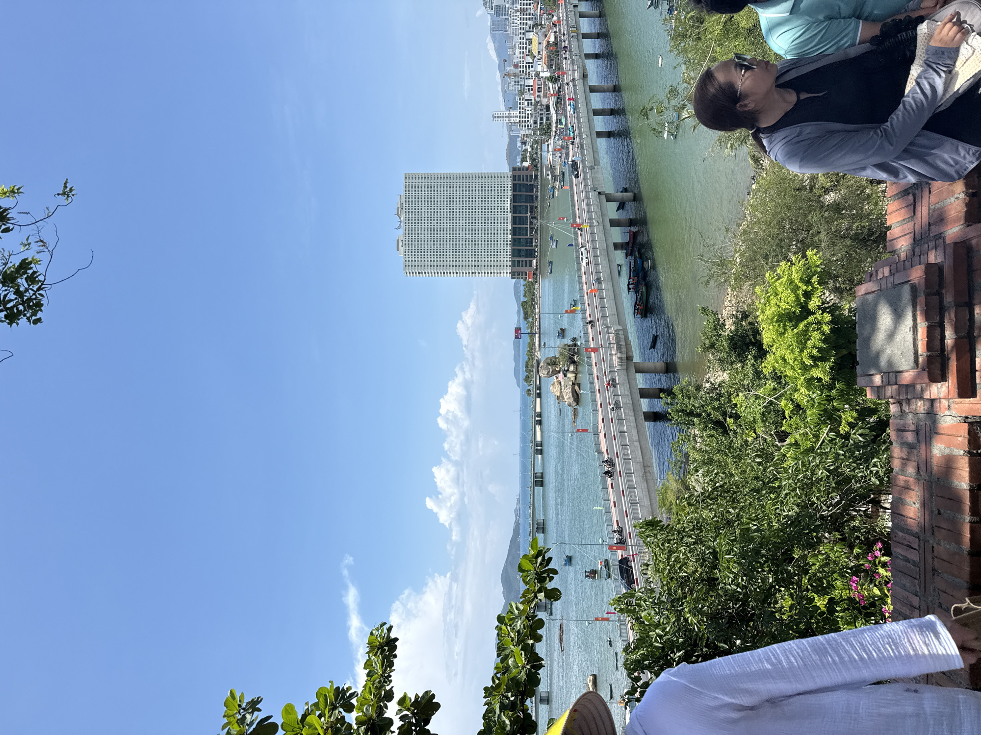 A vertical, eye-level shot shows a cityscape and bay in Bac Nha Trang Ward, Vietnam. A concrete bridge with red flags runs into the center of the image. Traffic is visible on the bridge. A large, grid-patterned building stands near the bridge. Many ships and boats are in the water. On the right, two people are looking out onto the view. A brick wall is behind them. Green plants and trees are in the foreground. A clear blue sky with a few clouds is visible. A portion of a straw hat and the sleeve of a white shirt are in the bottom of the image.