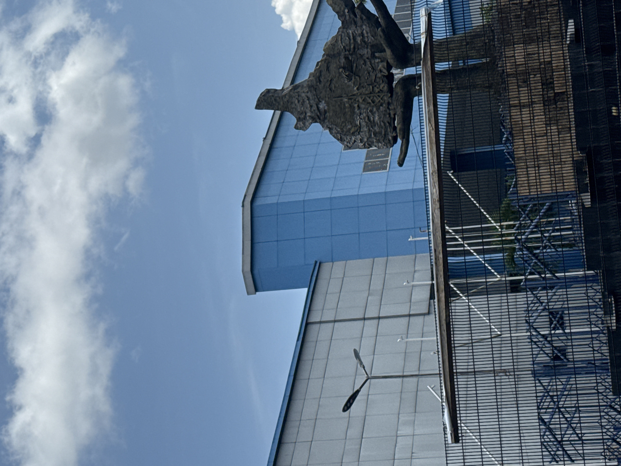The image depicts a low-angle view of buildings and a sculpture in Port Harcourt, Nigeria. The sky is blue with scattered white clouds. Two buildings are visible; one is light gray with a wind turbine mounted on its facade, and the other is blue. A dark-colored, textured sculpture appears in front of the blue building. A metal fence or scaffolding structure is in the foreground, partially obscuring the scene.