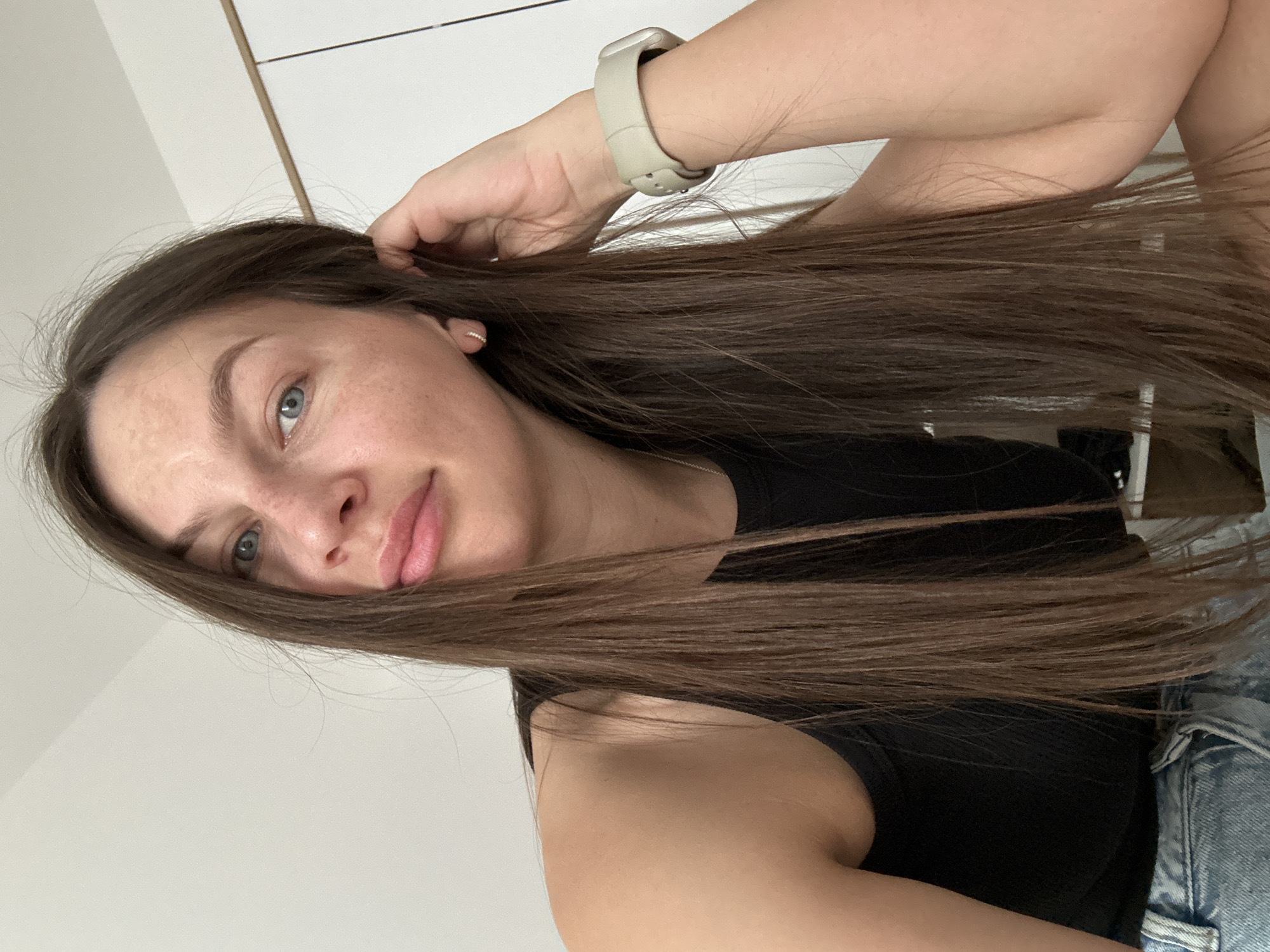 A selfie taken from a slightly elevated, angled perspective shows a fair-skinned female with long, straight brown hair. Her hair is parted to the side and cascades over her right shoulder. She has light blue eyes and is wearing a beige smartwatch on her left wrist. Her left hand is raised, fingers slightly curved, touching her hair near the ear. She is wearing a black tank top and appears to have denim clothing, likely jeans, near her waist. The background consists of a white wall with what appears to be a partially visible cabinet or door with light-colored trim. The photo appears to be taken indoors. There is no visible information to indicate the location in Budva, Montenegro.