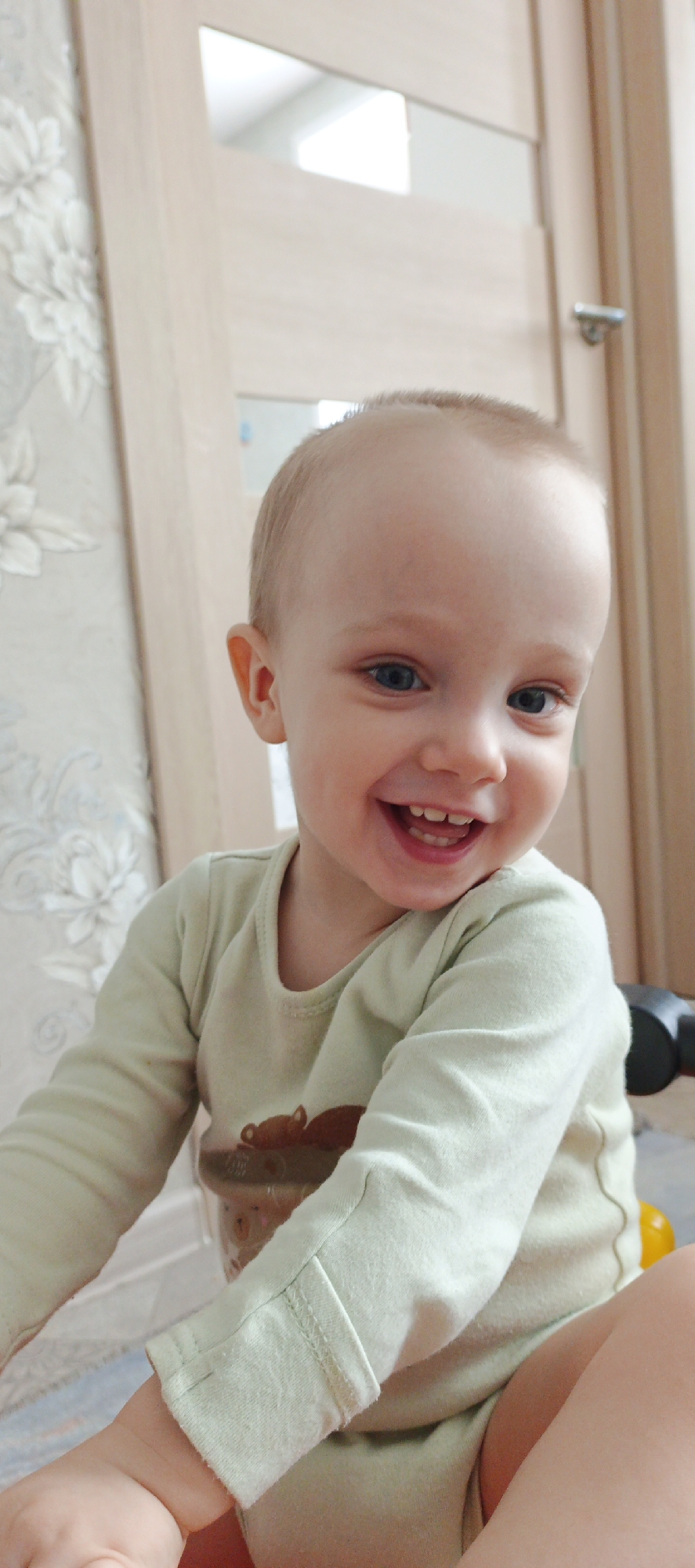 An indoor portrait in the city of Chaikovsky, Russia, features a young child, appearing to be a toddler, with short light hair and blue eyes, positioned centrally and looking directly at the camera. The child displays a wide, open-mouthed smile, revealing both upper and lower teeth. The child wears a long-sleeved, light green top with a brown graphic print resembling an animal on the chest. The lower right portion of the frame shows the child's bare leg and part of the arm, along with a dark, curved object and a yellow item partially visible in the background. The immediate background to the left consists of a wall covered in light-colored, patterned wallpaper featuring floral or botanical designs. To the right and behind the child is a light-toned wooden interior door or door frame, with a silver-colored handle visible on the right side. A bright, undefined reflection is present in an upper panel of the door structure.
