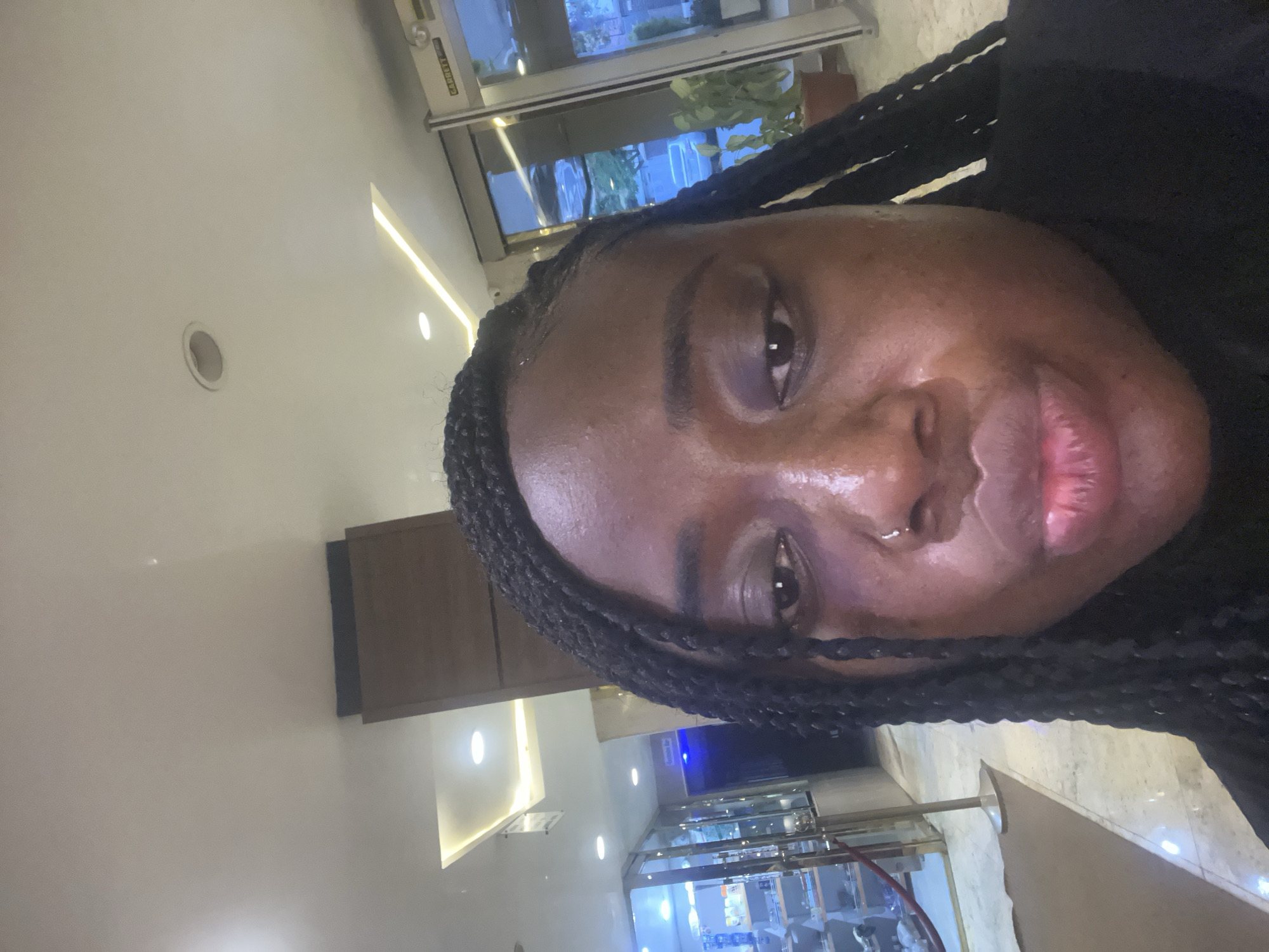 A selfie captures a person, likely female, with dark skin and braided hair, looking directly towards the camera. She has a nose piercing in her left nostril, and her lips are slightly parted with a neutral expression. She appears to be wearing a dark top.
The background indicates an indoor commercial or public setting, possibly a lobby or retail space. The ceiling features both circular recessed spotlights and linear strip lighting. Behind the person, a large glass window or partition reflects an outdoor scene showing green foliage and parts of buildings, suggesting daylight outside. A sign above the reflected area partially reads "GARRETT."
To the person's right, there are light-colored walls, some adorned with dark, rectangular, possibly wooden or metallic decorative panels. Further back, brightly lit display cases or counters are visible, emitting blue and white light, suggesting a retail environment with products. The floor is light-colored and reflective, contributing to the well-lit atmosphere. The image is taken in a portrait orientation but is rotated 90 degrees counter-clockwise. The setting implies a modern, contemporary space, potentially a secure entrance or a shop in Abuja, Nigeria.