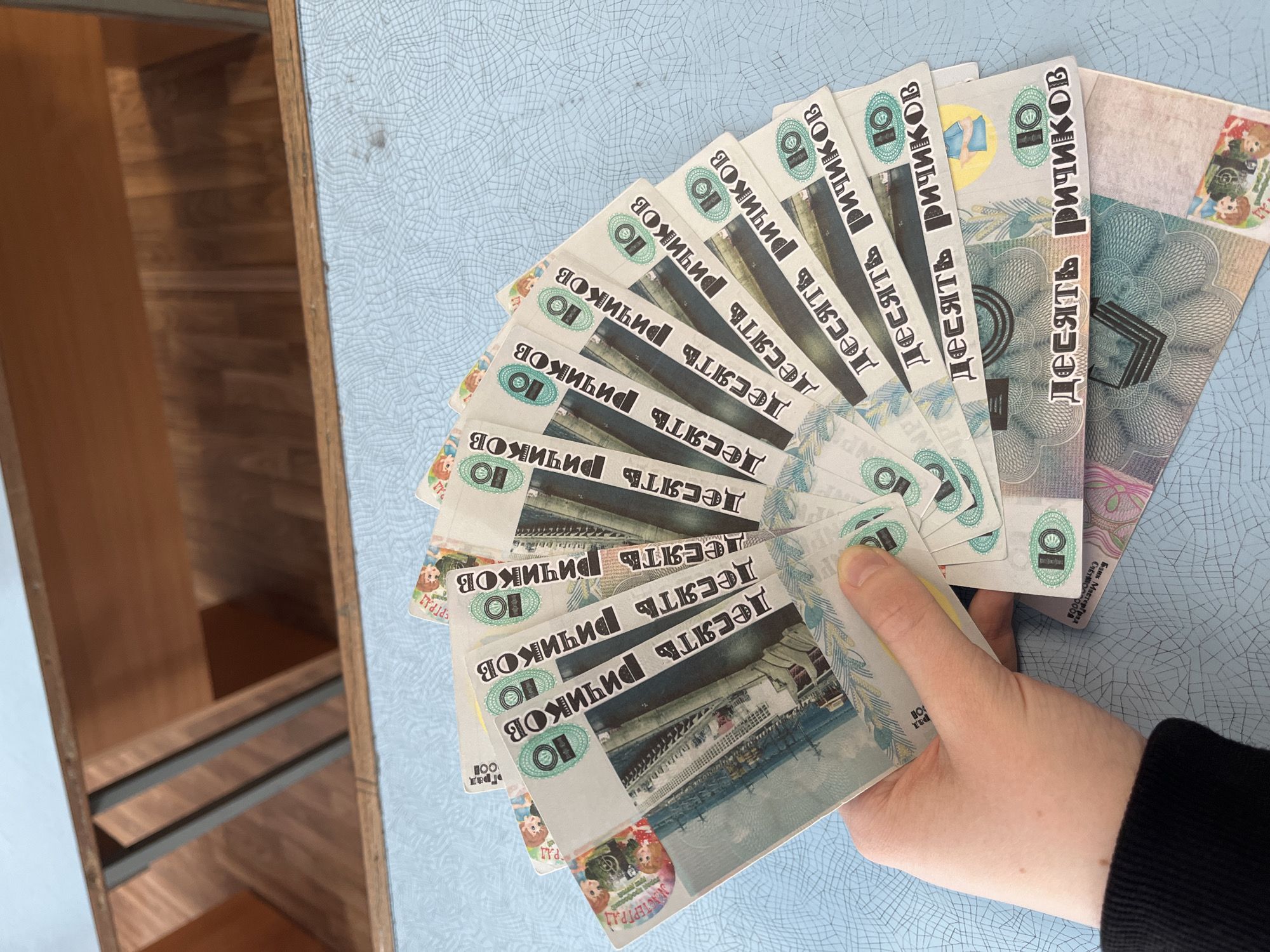 A hand holds a fan of "Ten Richikov" play money against a blue, cracked-pattern surface, with a partially visible wooden cabinet to the left. The play money features images resembling Russian currency, some with pictures of children and cityscapes. One bill includes "Bank Mastergrad" text. The words "Chaikovsky, Russia" do not appear in the image.