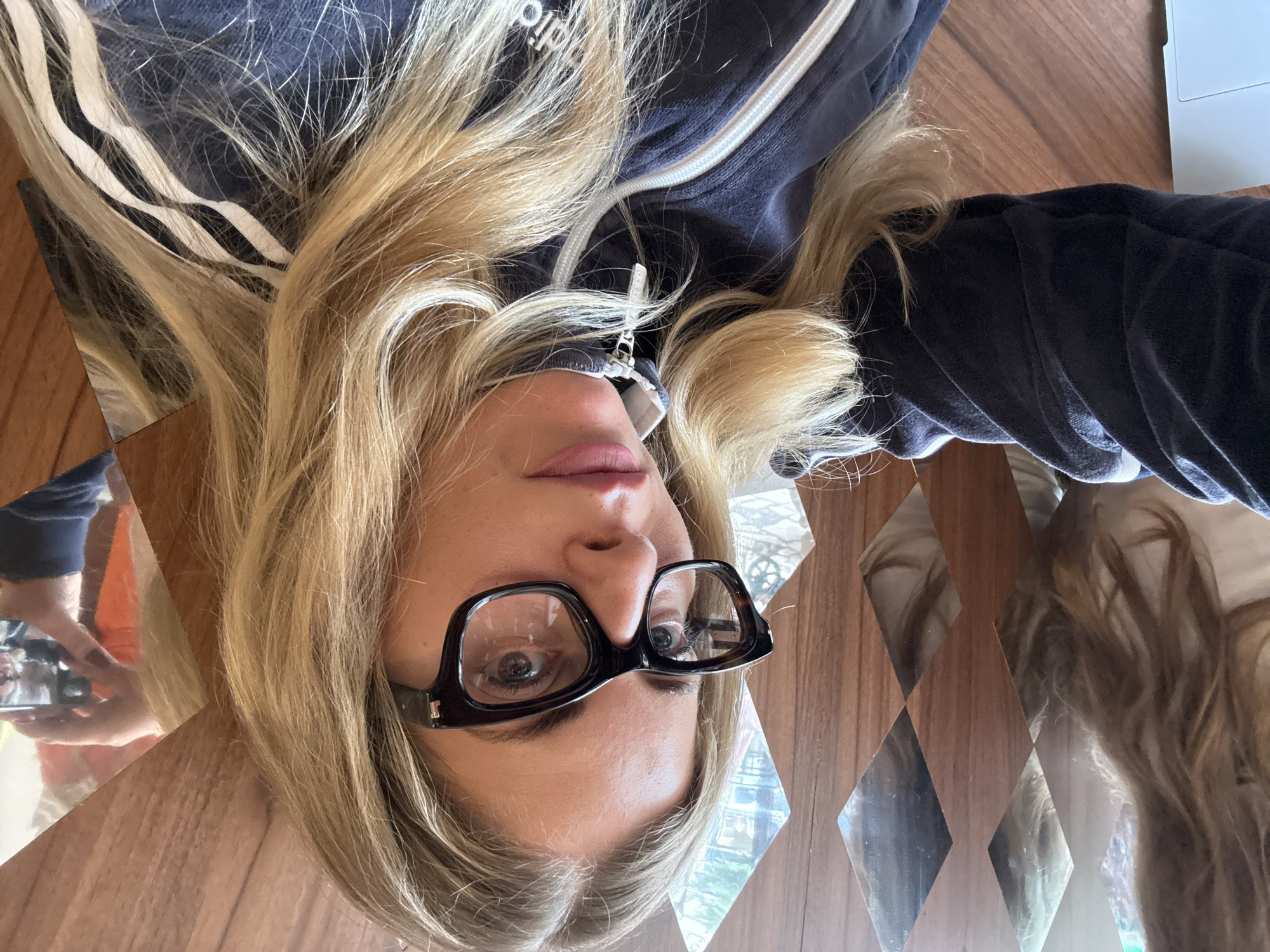 A fair-skinned woman with blonde hair is lying down with her head facing the camera. She is wearing black framed glasses. Her hair is spread out on a table, and she is wearing a navy blue Adidas track jacket. In the background, there is a series of diamond-shaped mirrors. The table appears to be made of wood. The location is not identifiable as Paris, France.