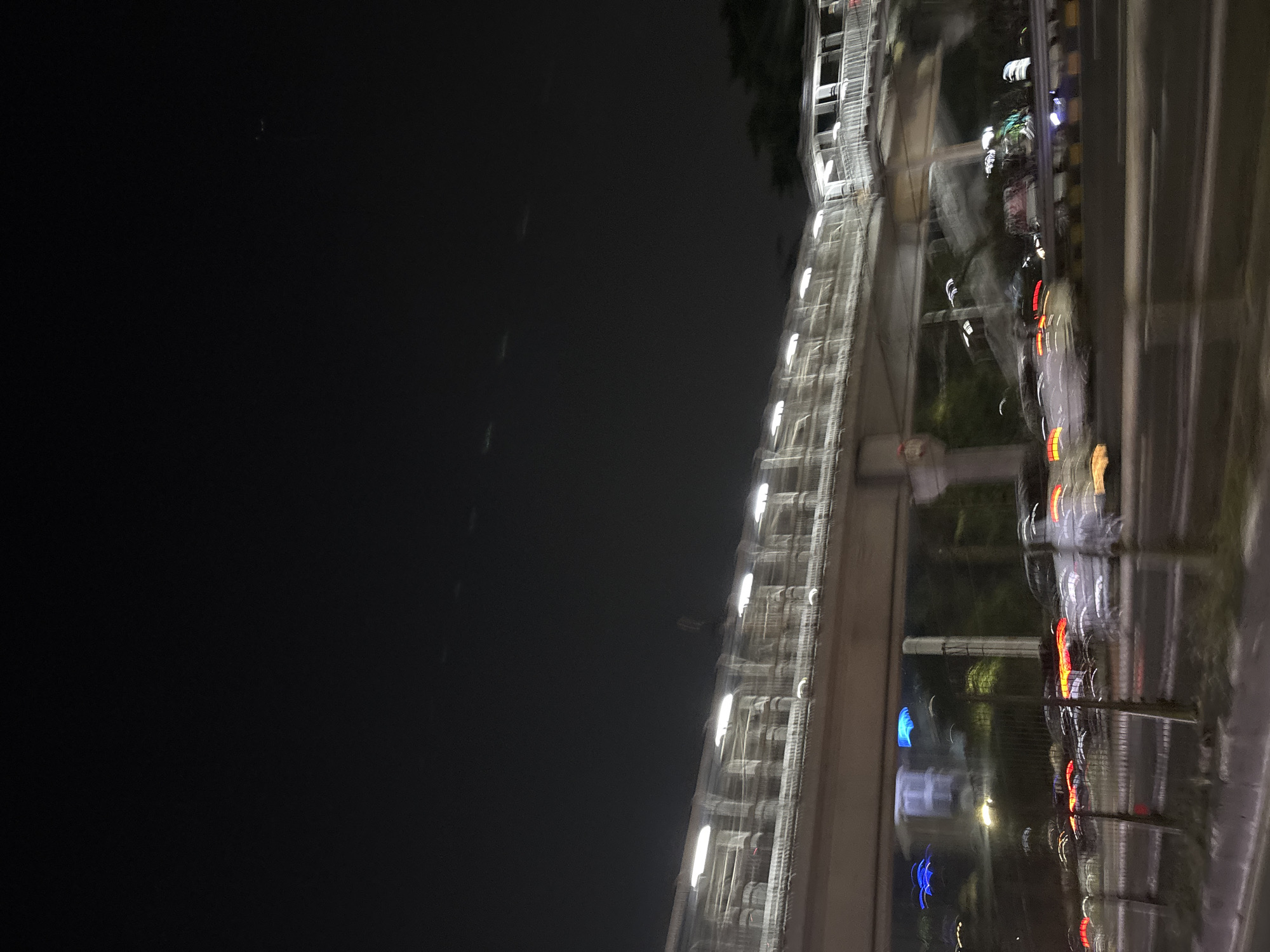 This is a night shot of a pedestrian bridge over a road in West Jakarta, Indonesia. The image is slightly blurred due to motion, giving a sense of movement. The pedestrian bridge has bright lights running along its length, casting a glow on the structure. Below the bridge, vehicles are visible on the road, with their headlights and taillights creating streaks of light. Some vehicles appear to be in traffic. Trees and buildings can be seen in the background, partially obscured by the darkness and motion blur. The sky is dark, indicating nighttime.