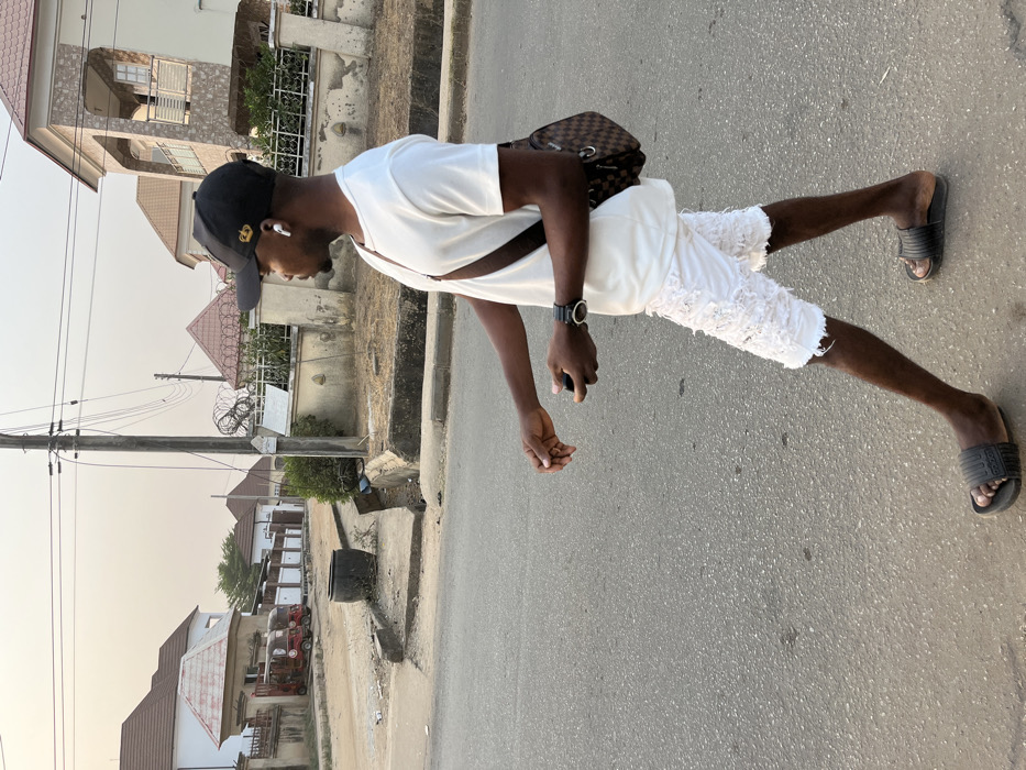 A man in a white t-shirt, tattered white shorts, and black slides stands on an asphalt road. He wears a black cap, a watch, and carries a checkered shoulder bag. He appears to be holding a phone or small object in his hand. Houses and power lines are visible in the background under a clear sky.