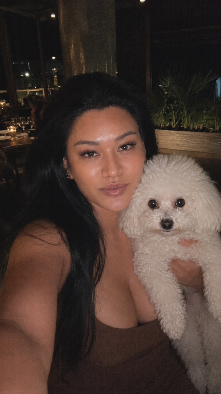 A woman is holding a white poodle in her arms, taking a selfie in a dimly lit, upscale restaurant with a city view at night. The woman has long, dark hair and is wearing a brown, ribbed dress. The poodle is fluffy and looking directly at the camera. In the background, there are tables with place settings and candles, as well as plants and a window overlooking a cityscape. There is no visible indication of Moscow, Russia in the image.