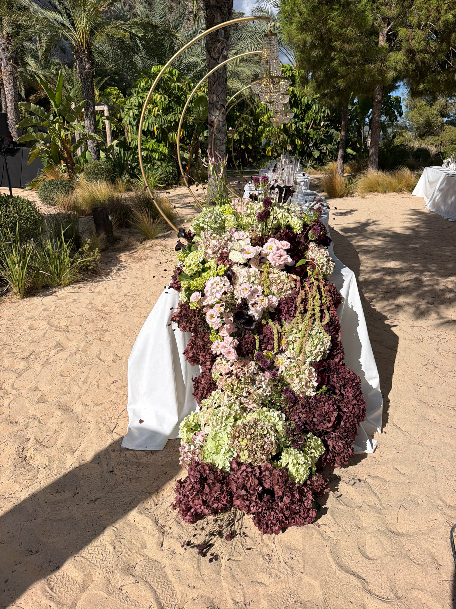 An outdoor wedding reception table is set on a sandy surface in Sant Josep de sa Talaia, Spain. The long table is draped with a white tablecloth and adorned with an abundant floral arrangement in shades of burgundy, pale pink, and light green. A large golden circular arch is positioned behind the table, with a crystal chandelier suspended from it. In the background, lush green trees and palm trees suggest a tropical or Mediterranean setting, with another white-draped table visible in the distance. The sandy ground shows tire tracks, indicating the area is accessible by vehicle. The scene is bathed in sunlight, casting shadows from the surrounding foliage onto the sand.