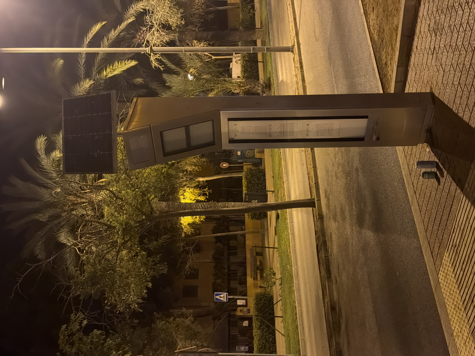 The image depicts a nighttime scene on a street in Calvià, Spain, likely near a bus stop. A modern, metallic bus stop shelter with a solar panel on top stands on the sidewalk. The shelter has a digital display panel and a printed schedule. Two cans are visible on the textured sidewalk in front of the shelter. Across the street, a road with faint lane markings is visible, bordered by a sidewalk and a strip of grass. Beyond the grass, a low hedge runs along the property line of buildings. One building appears to be a restaurant or shop with large windows and an awning. Palm trees and other trees are scattered throughout the scene, some illuminated by streetlights. A pedestrian crossing sign is visible on a pole near the building. The overall lighting suggests it is late evening or night, with artificial lights casting shadows.