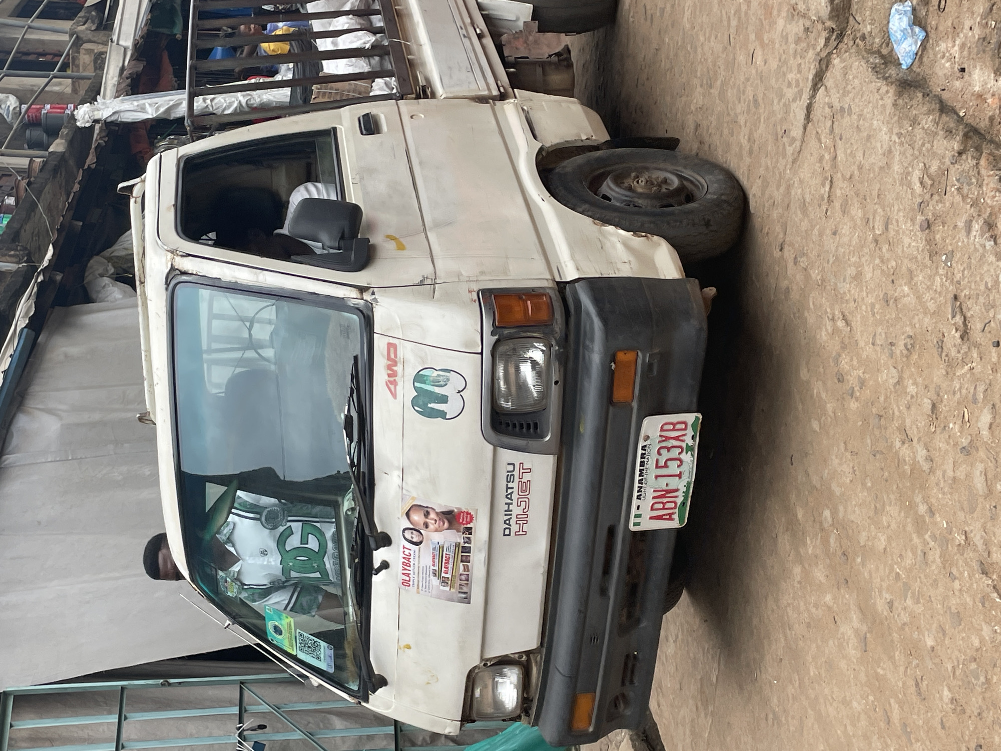 In Ogidi, Nigeria, a white Daihatsu Hijet pickup truck is parked on a dirt road. The truck's front bumper is damaged, and it has a license plate that reads "ABN-153XB" with "ANAMBRA" printed above it. A sticker on the truck's windshield displays a QR code and a green and white logo. Another sticker on the truck's hood promotes Olay Bact Triple Action Cream with images of women. A person wearing a white and green shirt with a "DG" logo is visible through the windshield. The truck's bed is filled with white sacks. In the background, there are stalls with corrugated metal roofs and various items stacked, suggesting a market or commercial area. The environment appears weathered and somewhat unkempt, with loose debris on the ground.