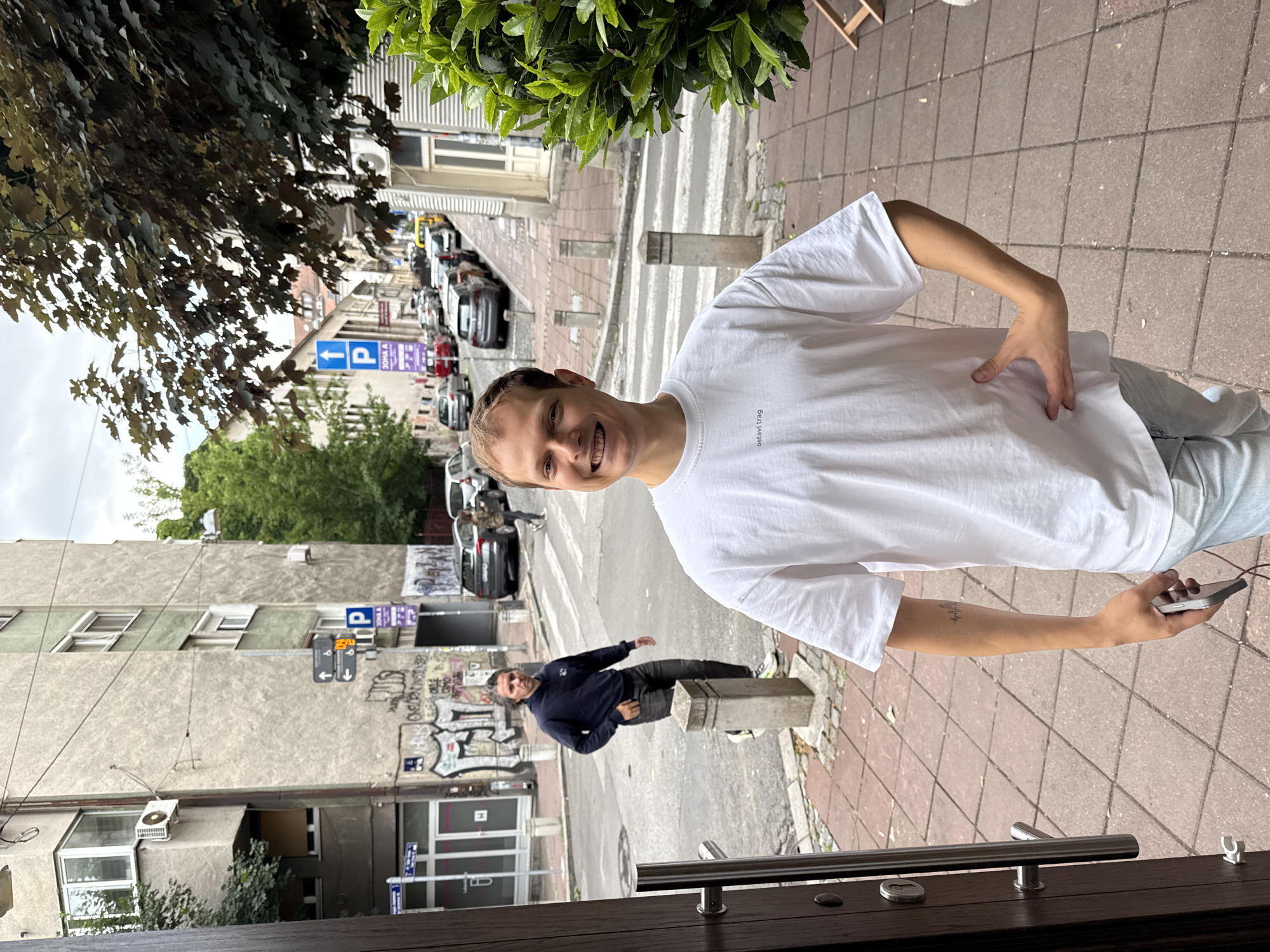 A young man with light brown hair and a white t-shirt stands on a sidewalk in Belgrade, Serbia, smiling at the camera. He is holding a phone in his right hand and his left hand is on his hip. In the background, a man in a dark blue hoodie and jeans walks across a pedestrian crossing. The street is lined with buildings, parked cars, and trees. A parking sign with an accessibility symbol is visible on a pole. The sky is overcast.