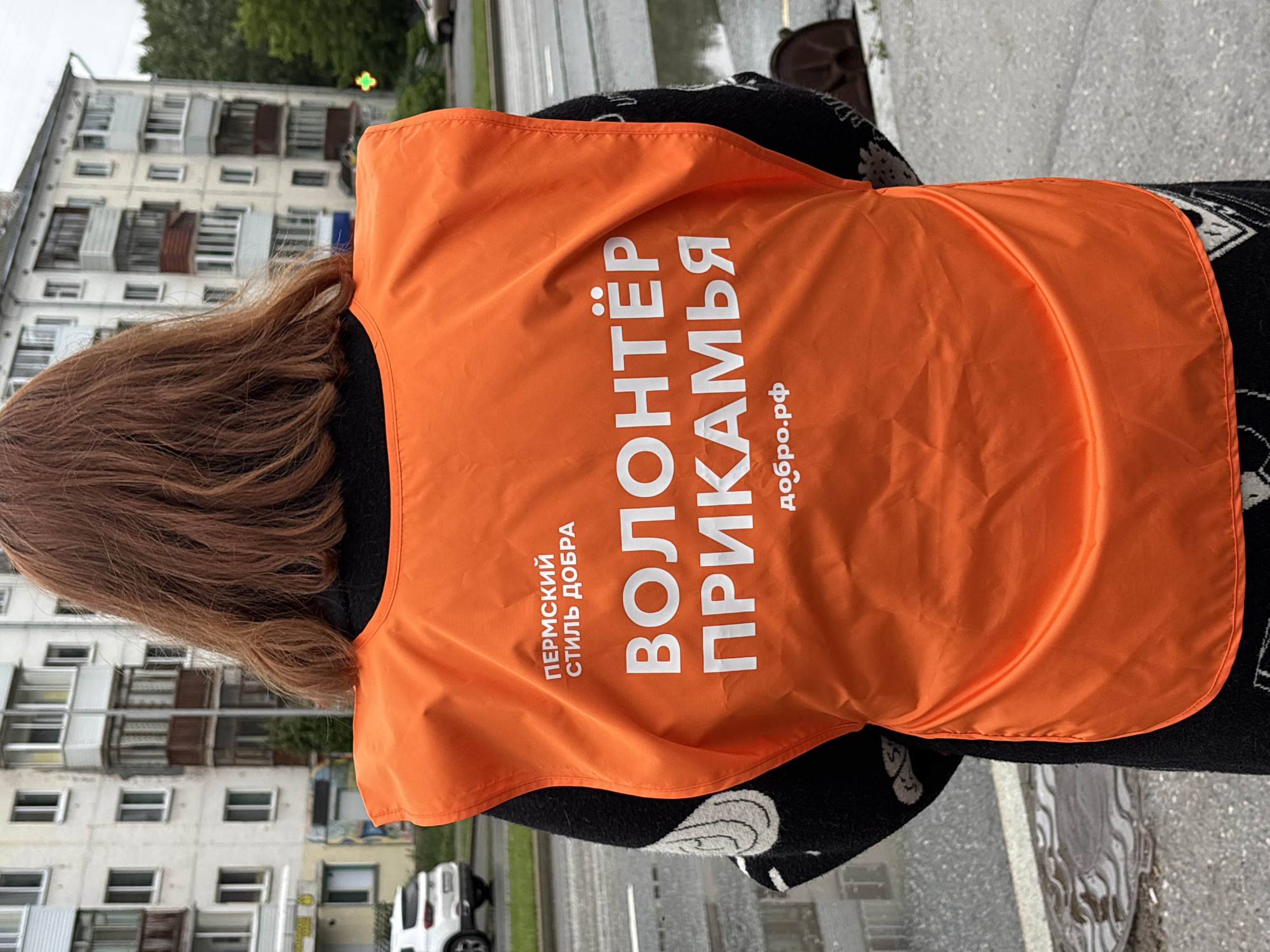 An individual is depicted from the back, centrally positioned, wearing an orange sleeveless vest over a dark long-sleeved garment patterned with white or light-colored designs. The person has reddish-brown hair reaching to the shoulders. The orange vest features white Cyrillic text: "ПЕРМСКИЙ СТИЛЬ ДОБРА" (Permian Style of Goodness) at the top left, "ВОЛОНТЁР ПРИКАМЬЯ" (Volunteer of Prikamye) prominently in the center, and "добро.рф" (good.rf) at the bottom. Prikamye is the region surrounding the Kama River, which includes the city of Perm, Russia.
The scene is an urban outdoor environment with a wet asphalt road and sidewalks, suggesting recent precipitation. To the immediate left of the individual, a reflective round metal manhole cover is visible on the wet ground. In the background, multiple light-colored multi-story residential buildings with numerous windows and balconies line the street. On the left side of the street, a white SUV is parked or moving slowly. On the right side, a green illuminated cross, indicative of a pharmacy, is visible near one of the buildings. A partial view of a brown or beige sedan is also present on the road to the right. Sparse greenery and trees are interspersed between the buildings and the street. The lighting is diffuse, consistent with an overcast daytime sky.