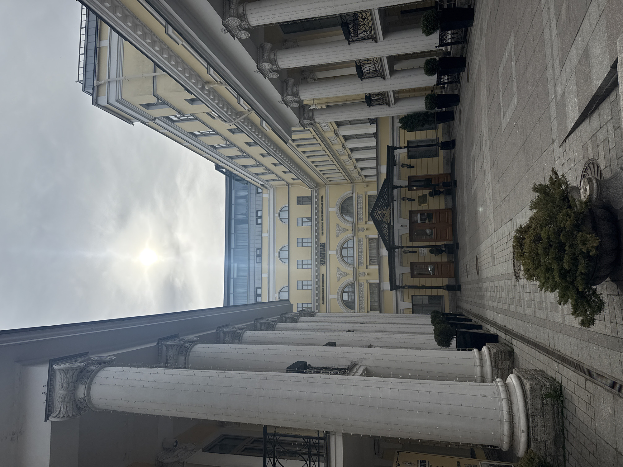 Captured from a low-angle perspective in Saint Petersburg, Russia, the image features a courtyard enclosed by classical architecture. The buildings are light yellow with white column-lined facades, decorated with strings of lights. The sky is overcast, with the sun a bright spot piercing through the clouds. The ground is paved with gray stones, lined with hedges and potted plants.