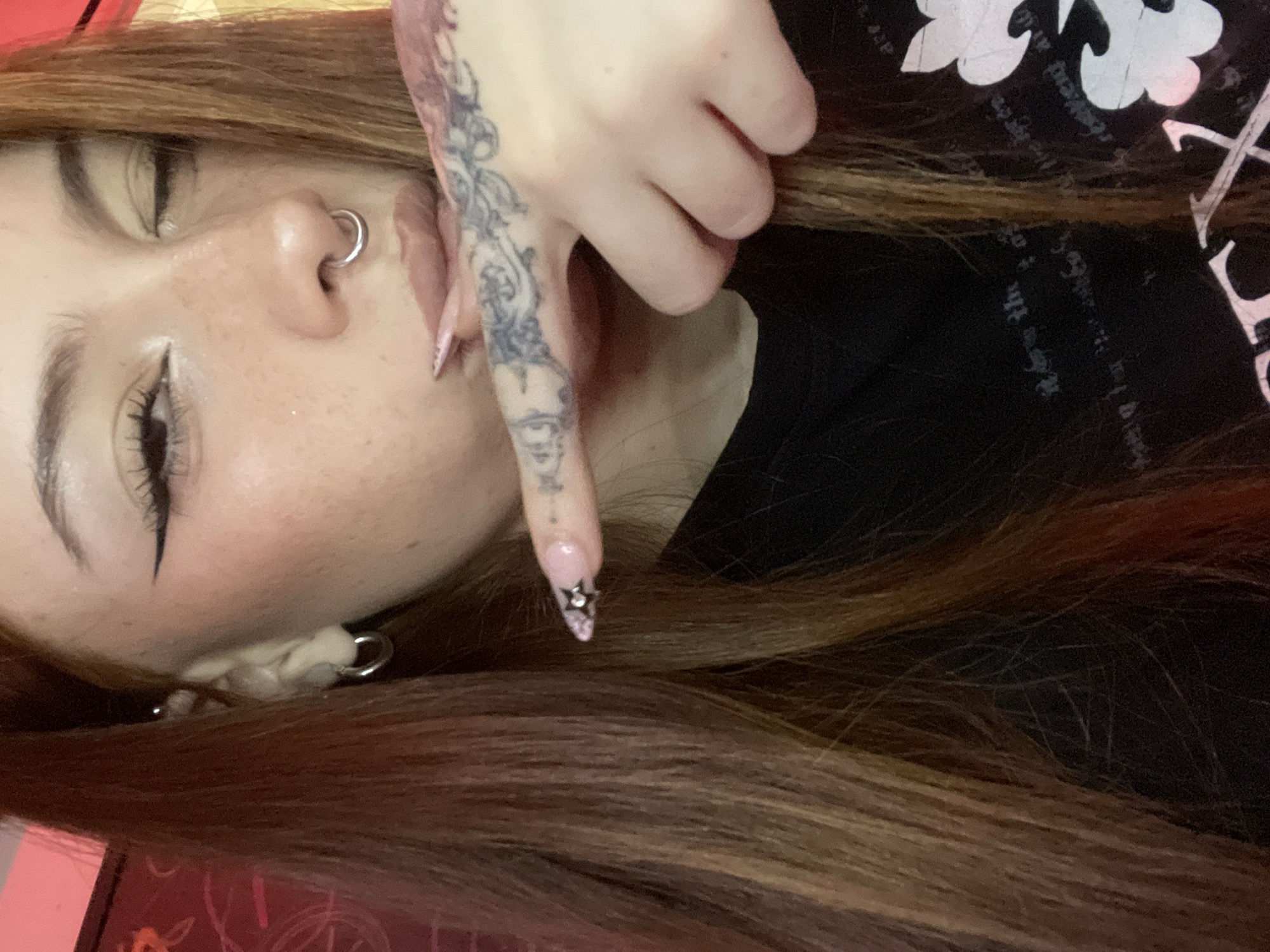 The image depicts a close-up of a person with fair skin, brown hair, and a nose ring. The person's left eye is visible, featuring dark eyeliner and eyelashes. A hand with a detailed tattoo on the index finger is positioned near the person's face. The fingernail of the tattooed finger is decorated with a small, star-shaped embellishment. The person is wearing a black shirt. The location of Buenos Aires, Argentina is not evident from the image.