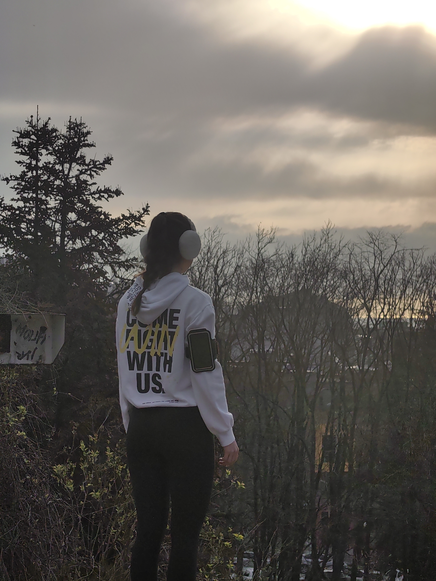 A medium outdoor shot depicts a woman standing with her back to the camera, wearing a white hoodie with yellow and black lettering, black leggings, and headphones. Her phone is strapped to her arm. She is looking out at the horizon. In the background are barren trees and the sky. The image was taken in Varna, Bulgaria.