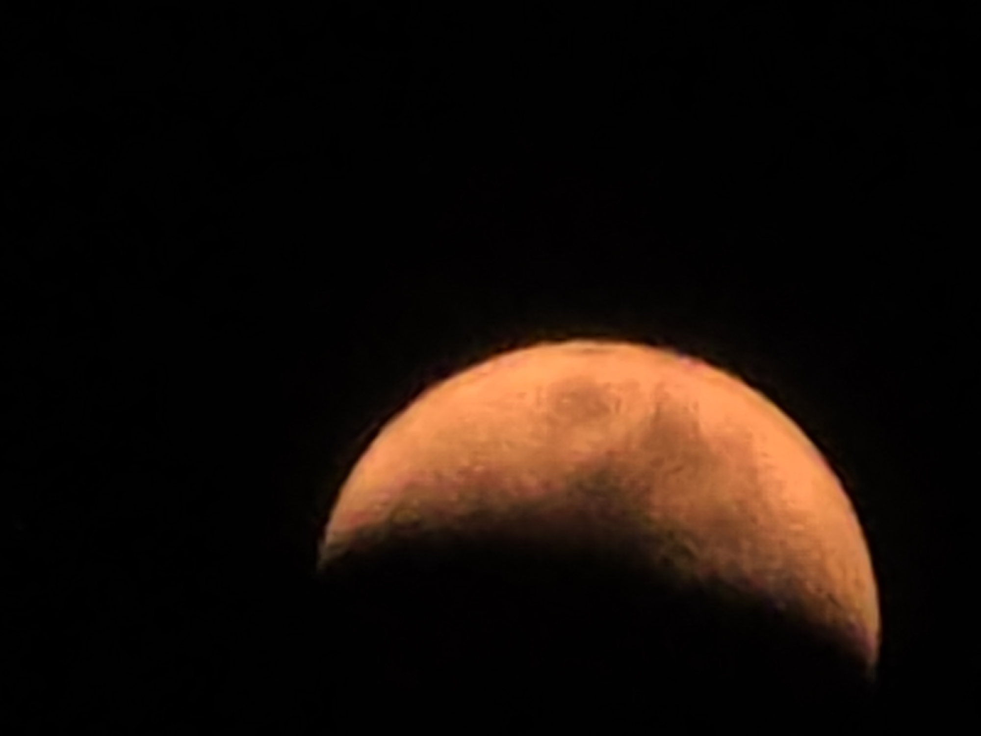 The image is a close-up of a celestial body, likely the moon, against a black background. The illuminated portion of the object is orange-yellow and displays textured features, suggesting craters or other surface variations. The left side of the object is dark, indicating it is not fully illuminated. The image is cropped tightly around the object, with the majority of the frame filled with the dark background. There is no discernible urban environment, city, or specific location such as Yambol, Bulgaria, visible in this image.