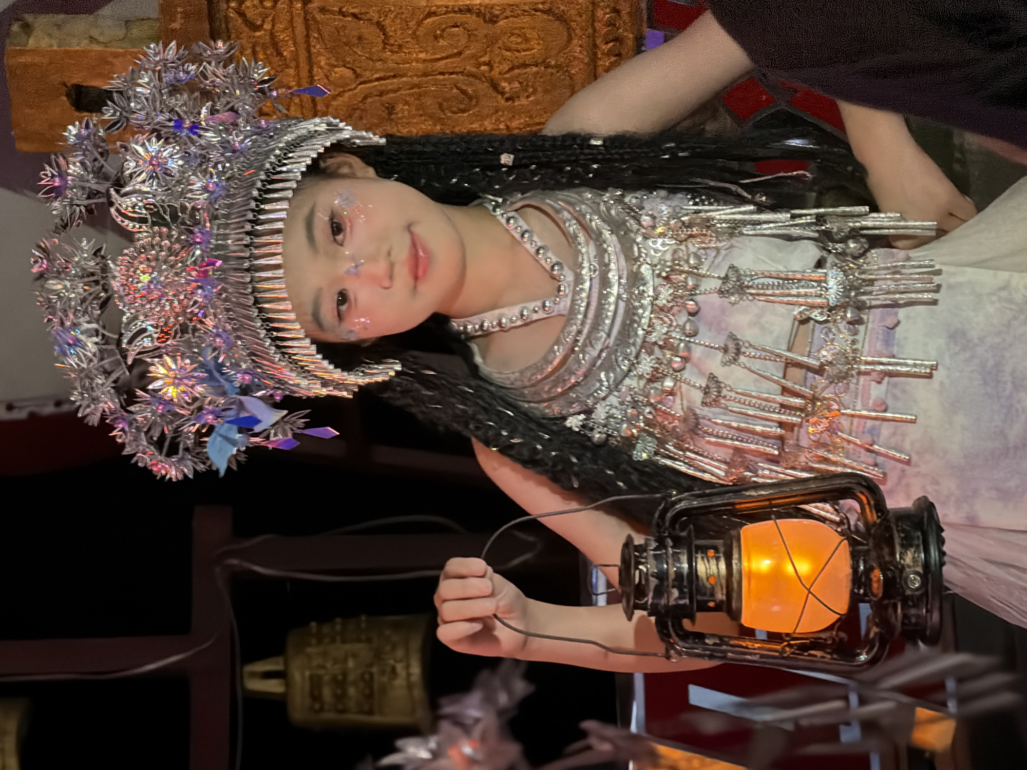 The image depicts a young East Asian girl dressed in traditional attire, holding an oil lantern. She wears an elaborate silver headdress adorned with crystals and decorative elements, along with a silver necklace and ornate chest piece. Her hair is styled in long braids, and she has subtle glitter makeup on her face. The background is dark, with hints of gold bells and what appears to be a decorative structure. The overall scene suggests a cultural performance or festival, potentially in the Yuzhong District of China, known for its rich cultural heritage.