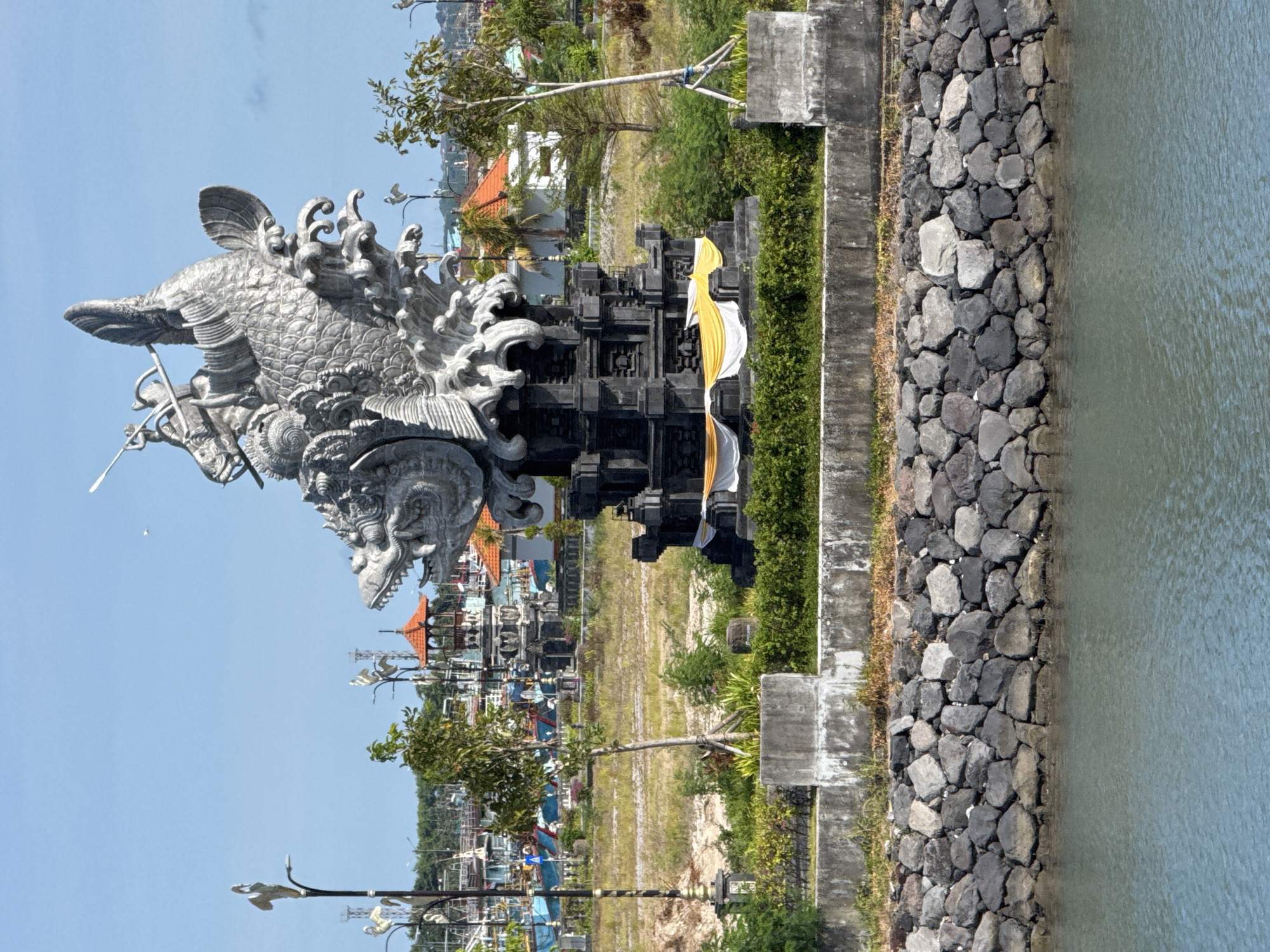 This image captures a scene in Denpasar, Indonesia. The primary subject is an elaborate stone statue of a creature resembling a fish or dragon, adorned with ornate carvings. This statue is positioned atop a dark-colored, multi-tiered structure that appears to be a Balinese gate or shrine, with a white and gold cloth draped across it.
The scene extends to reveal a relatively flat, grassy area dotted with small trees and structures resembling Balinese shrines or pavilions. In the background, there are glimpses of buildings with orange roofs and what appear to be fishing boats in the distance.
To the right of the statue, a stone wall made of large, dark-colored rocks leads down to a body of water, possibly a canal or the sea. The sky is a clear, light blue, suggesting a sunny day. The image is oriented vertically, giving a sense of the vertical scale of the statue and the surrounding landscape.