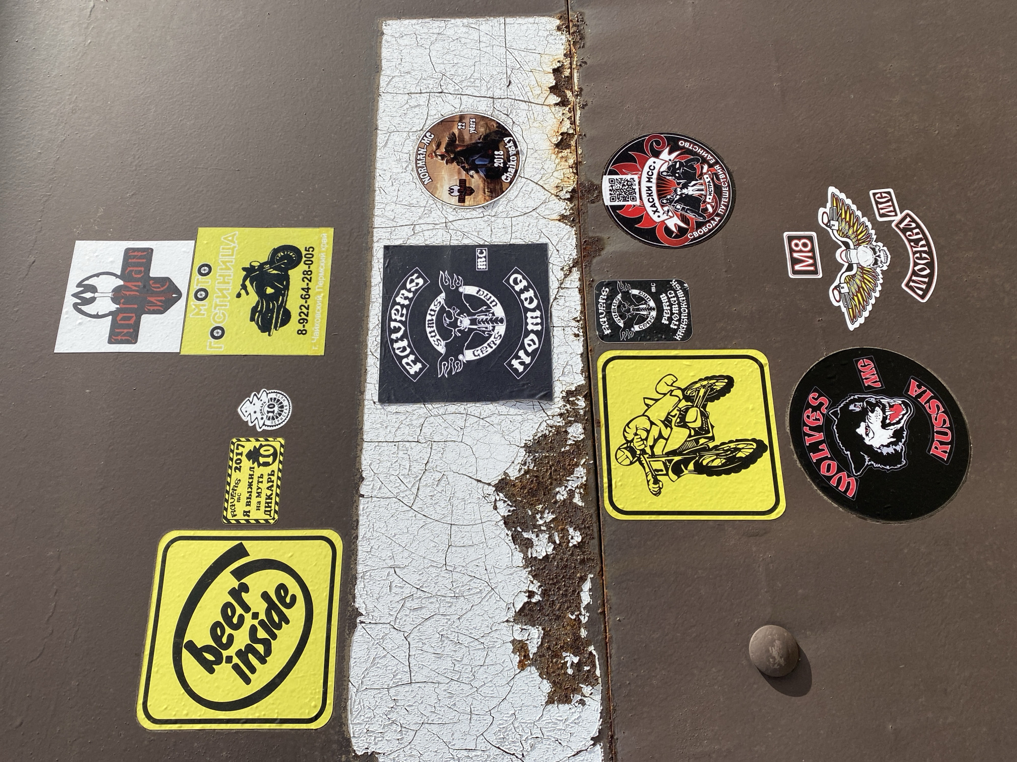The image shows a collection of stickers affixed to a brown, possibly metal, surface with a vertical strip of cracked white paint and rust running down the center. The stickers have motorcycle club insignia from "Nomad MC" and "Ravens MC", and a motorcycle-themed sticker advertising "Moto Gostinitsa" in Chaikovsky, Perm Krai with a phone number. Other stickers include "Beer Inside", "Wolves MG Russia", "M8" with an eagle and skull, "Moskva MG", "Khaski MCC", "Norman MC" and "I survived on Dikary 10". A small, round rivet is visible towards the bottom right.