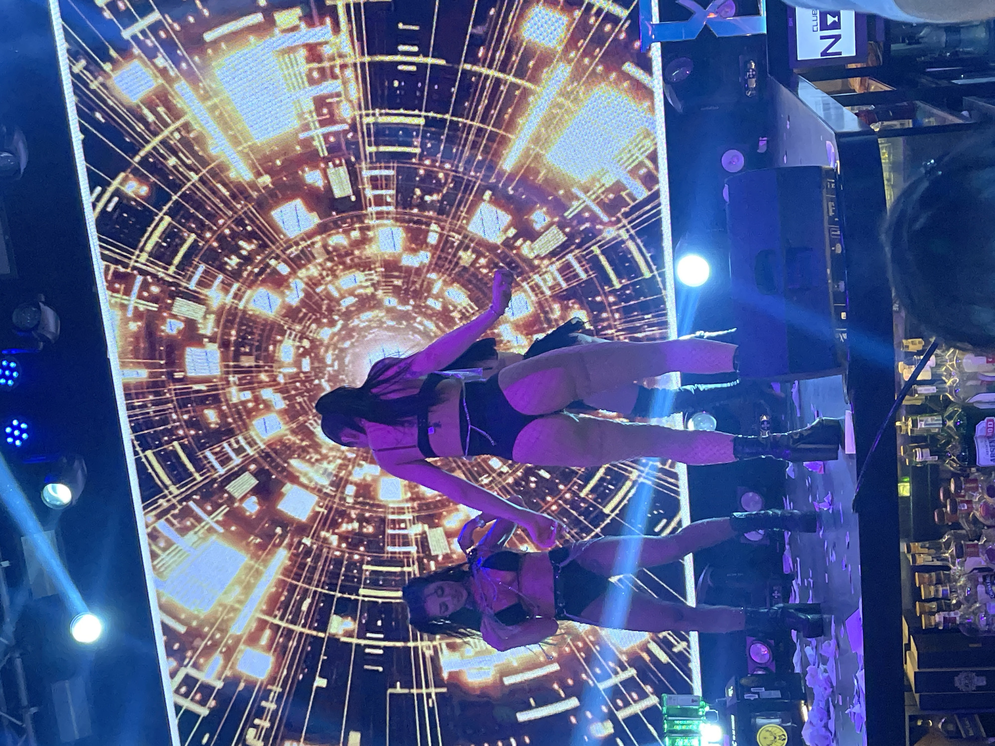 The image depicts two women in minimal clothing standing on a stage in what appears to be a nightclub. The background features a large LED screen displaying an abstract geometric pattern. The women are wearing dark-colored attire and boots. The environment includes stage lighting and elements of a bar or club setting. There is a sign that says "Club Next", so the scene could be Kazanlak, Bulgaria.