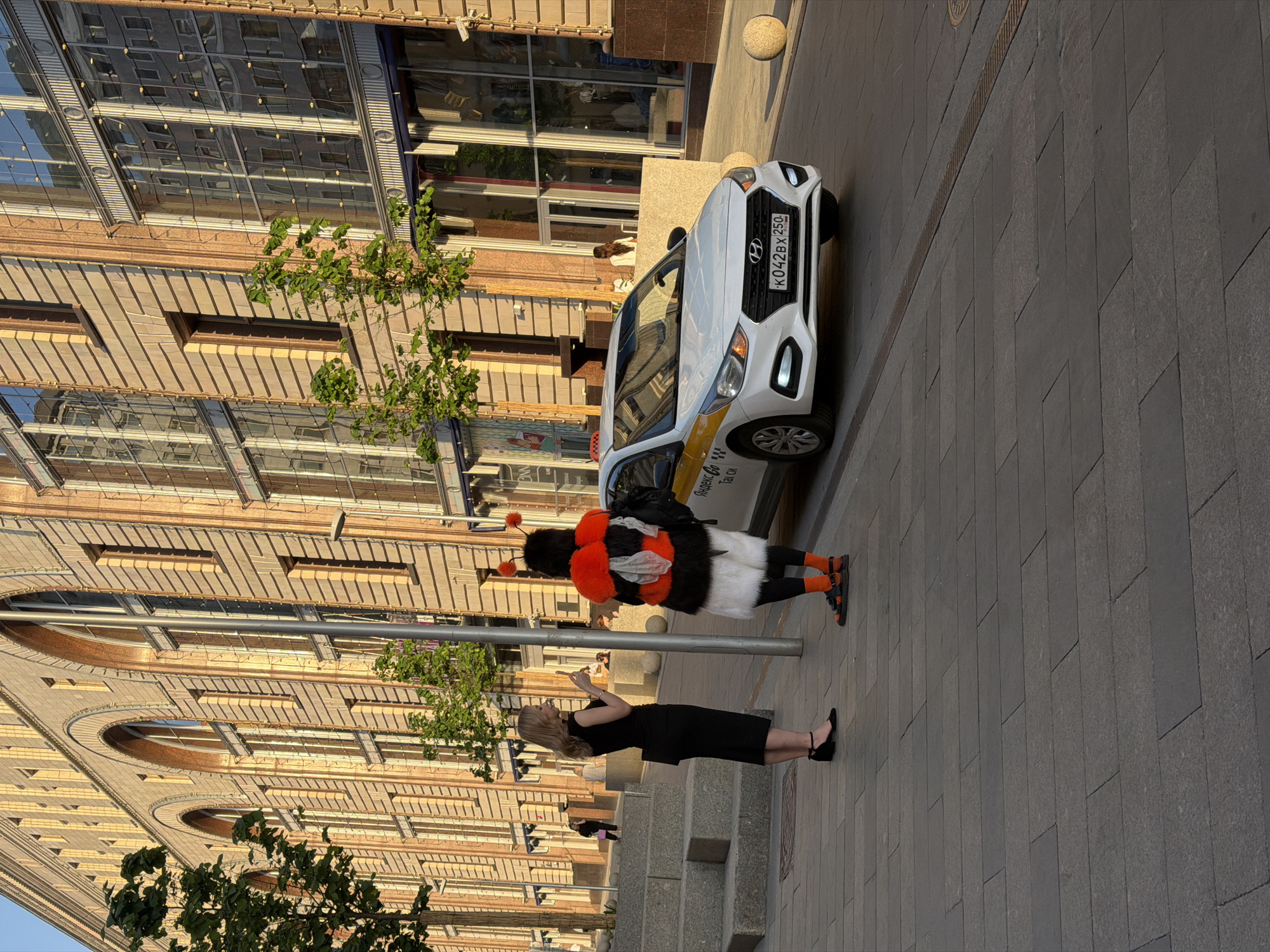 A woman in a black dress stands on a sidewalk in Moscow, Russia, facing a person dressed in a furry bumblebee costume. The woman appears to be speaking or gesturing towards the costumed person. Behind the bee costume, a white taxi is parked. The license plate of the taxi is visible, reading "K042BX 250". The backdrop consists of a large building with architectural details, including arched windows and multiple stories, suggesting a European-style city street. The photo is taken in daylight, with shadows indicating a time of day when the sun is at an angle. The sidewalk is paved with grey stone bricks.