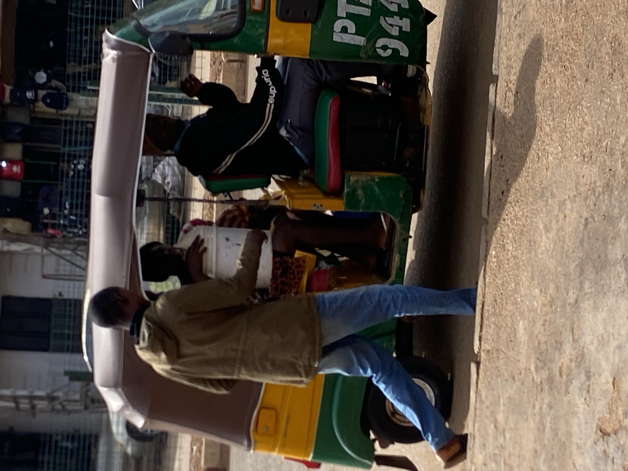 The image, oriented vertically, captures a daytime street scene in Bukuru, Nigeria, featuring a three-wheeled auto-rickshaw and several individuals.
The central subject is a green and yellow auto-rickshaw, commonly known as a 'Keke Napep', parked or moving slowly on a light-colored ground surface. The side of the auto-rickshaw's roof clearly displays white text: "PTA 944". Inside, a driver, wearing a dark jacket with "supremo" visible on the sleeve, is seated behind the handlebars. In the rear passenger section, another person is visible, holding a white bucket or container, with their legs extended.
Standing on the right side of the auto-rickshaw, facing it, is a man wearing a light brown or tan jacket, blue jeans, and brown sandals or slippers. He appears to be either interacting with the vehicle, waiting to board, or having just alighted.
The setting suggests an urban or semi-urban environment. The background shows structures that resemble market stalls or buildings with metal grates, hinting at a commercial area. Bright sunshine is evident from the distinct shadows cast on the ground to the right of the auto-rickshaw and the standing man. The overall scene conveys typical daily activity on a street in Bukuru, Nigeria.