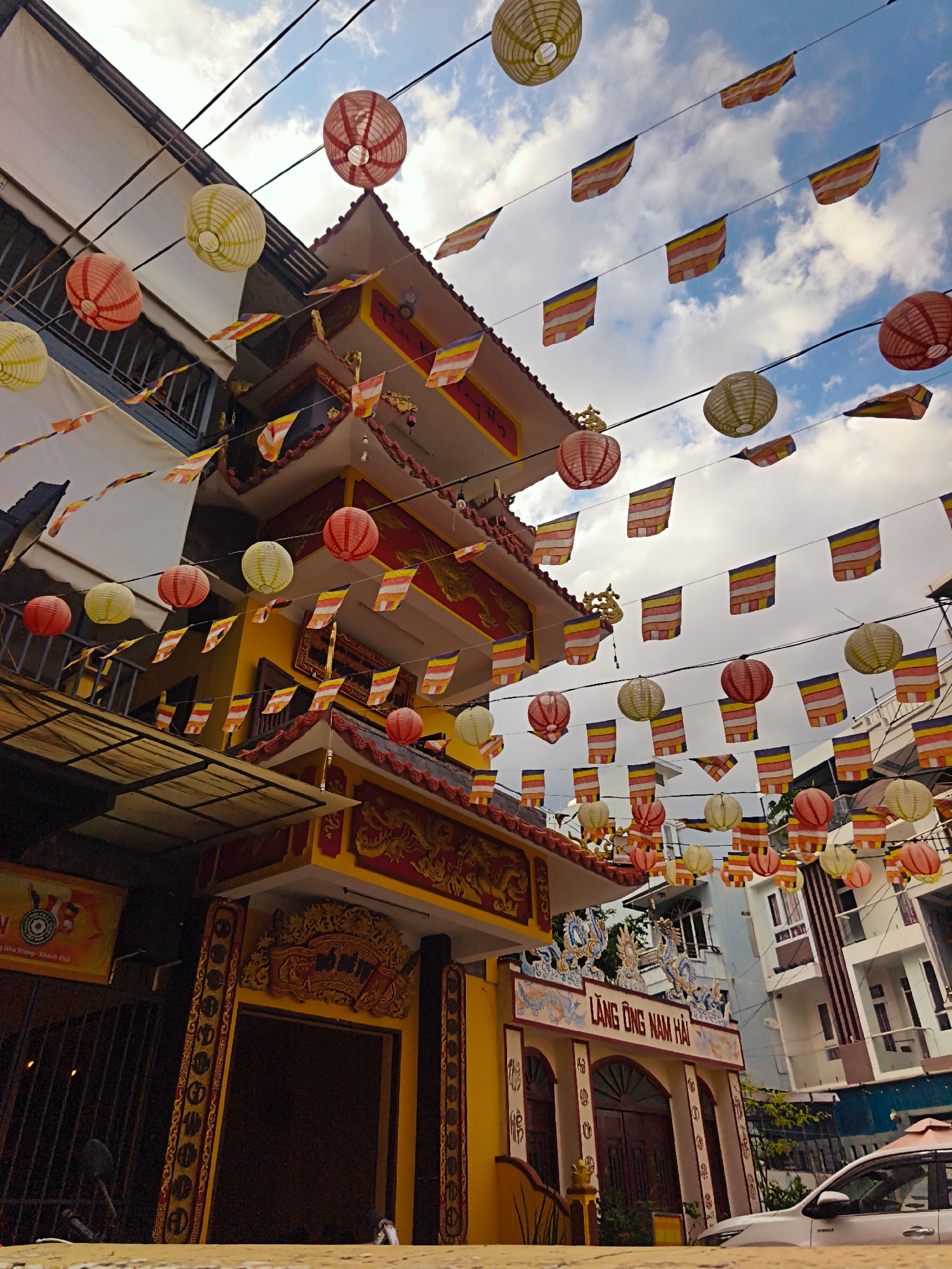 A low-angle, wide shot captures a Buddhist temple adorned with numerous colorful paper lanterns and prayer flags strung across wires. The temple, a multi-tiered structure with a predominantly yellow exterior and red tiled roofs, is decorated with ornate golden dragon motifs. In front of the temple, a sign with Vietnamese text reads "LĂNG ÔNG NAM HẢI." The temple is situated on a street in Nha Trang, Vietnam. The sky above is partly cloudy, with a mix of blue sky and white clouds. The foreground shows a low concrete wall and a white car parked on the right. The scene is viewed from street level, looking up towards the temple and the sky.